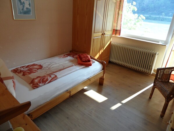 A single bed with a floral bedspread, wooden furniture, a wicker chair, and a view of the outdoors through a window. - Hotel Rheinkönig (Kamp-Bornhofen)