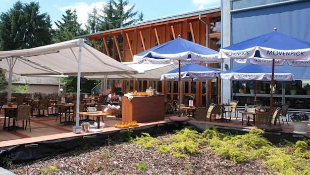 Un área de comedor al aire libre con mesas y sillas, cubierta por un dosel blanco y sombrillas azules, y una estructura de madera al fondo.