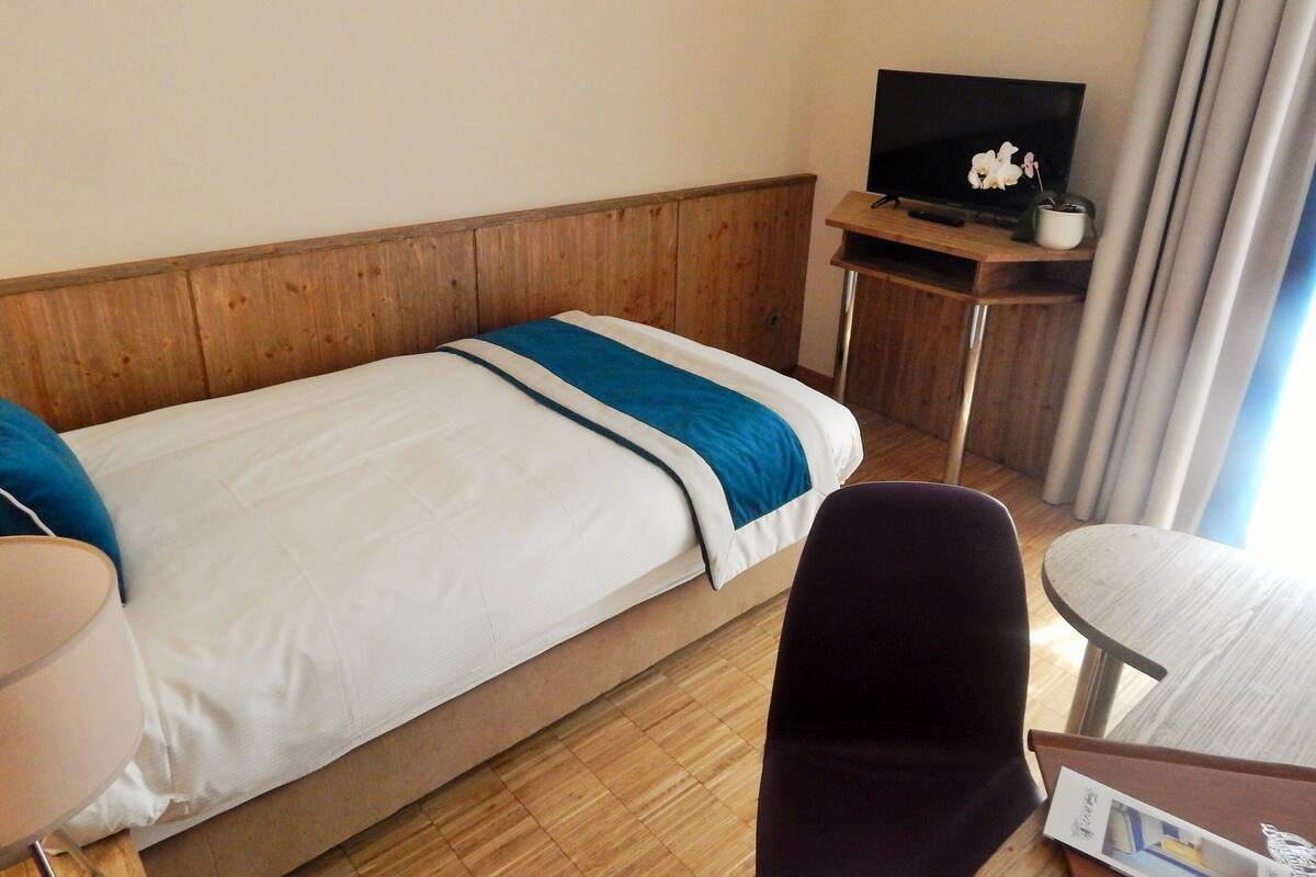 Habitación de hotel con una cama individual, un televisor sobre un atril de madera, una mesita pequeña y una silla.