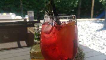 Un vaso de bebida roja con una ramita de romero sobre una mesa blanca. Se ve una botella de Fuze Tea al fondo.