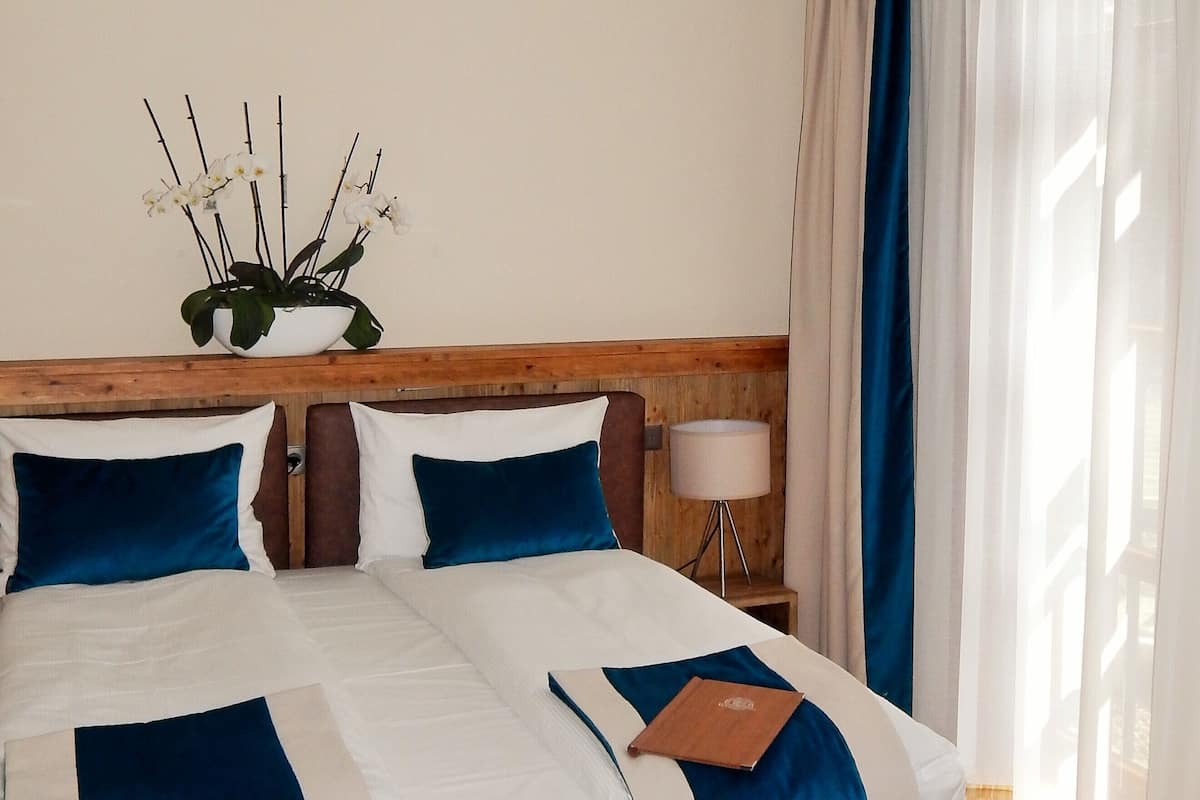 Una cama doble con cabecera de madera, ropa de cama blanca y azul, una lámpara de noche y una planta en maceta.