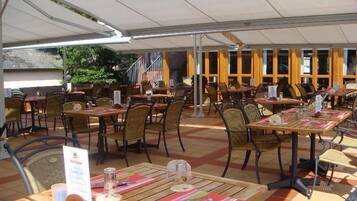 Un café al aire libre con mesas y sillas de madera, cubierto por un toldo blanco.