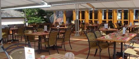 An outdoor cafe with wooden tables and chairs, covered by a white canopy.