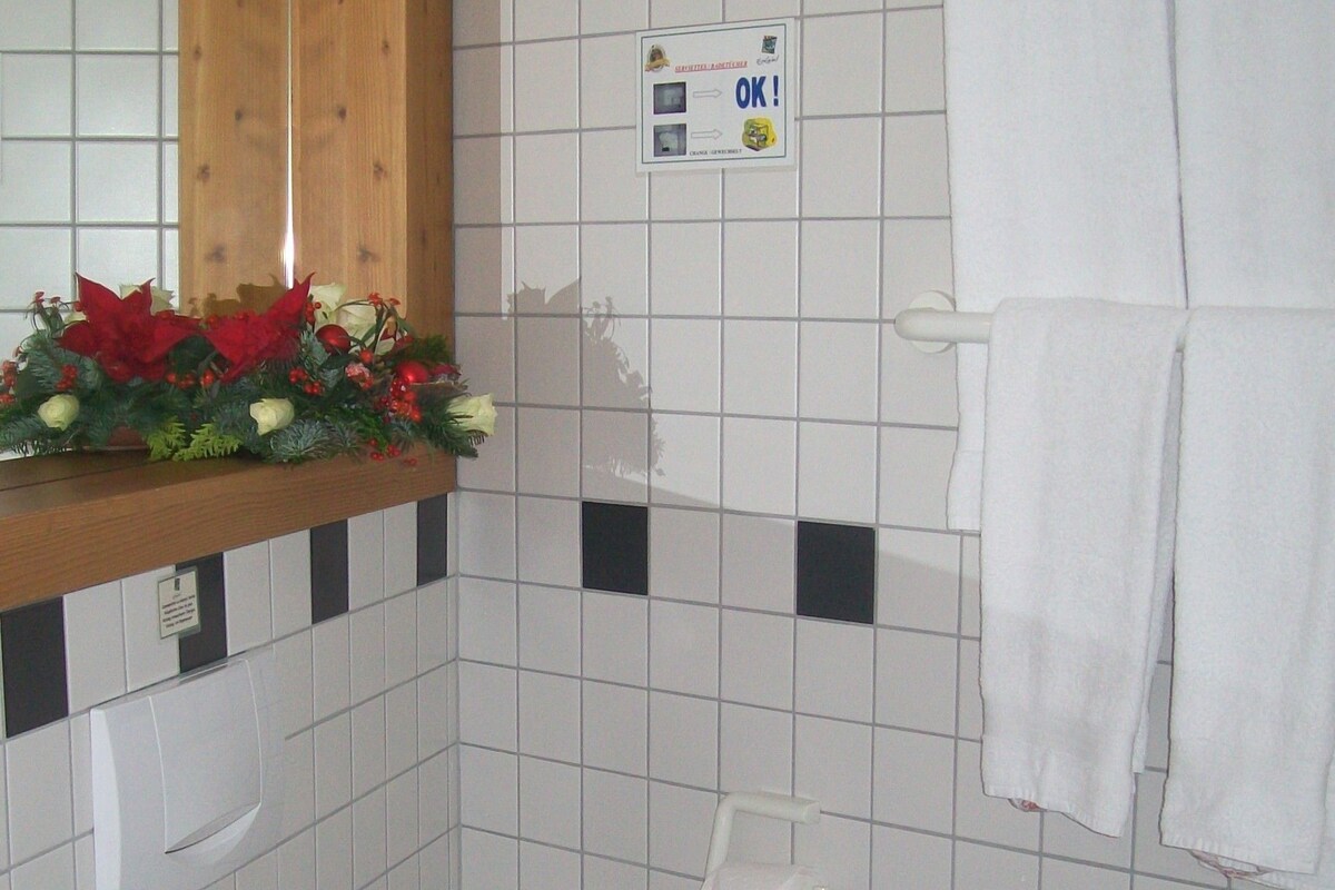 Un baño con inodoro, lavamanos y espejo sobre el lavamanos. Hay un letrero en la pared y un perchero con toallas.