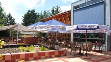An outdoor patio area with multiple tables and chairs, covered by umbrellas, and a building with large windows in the background.