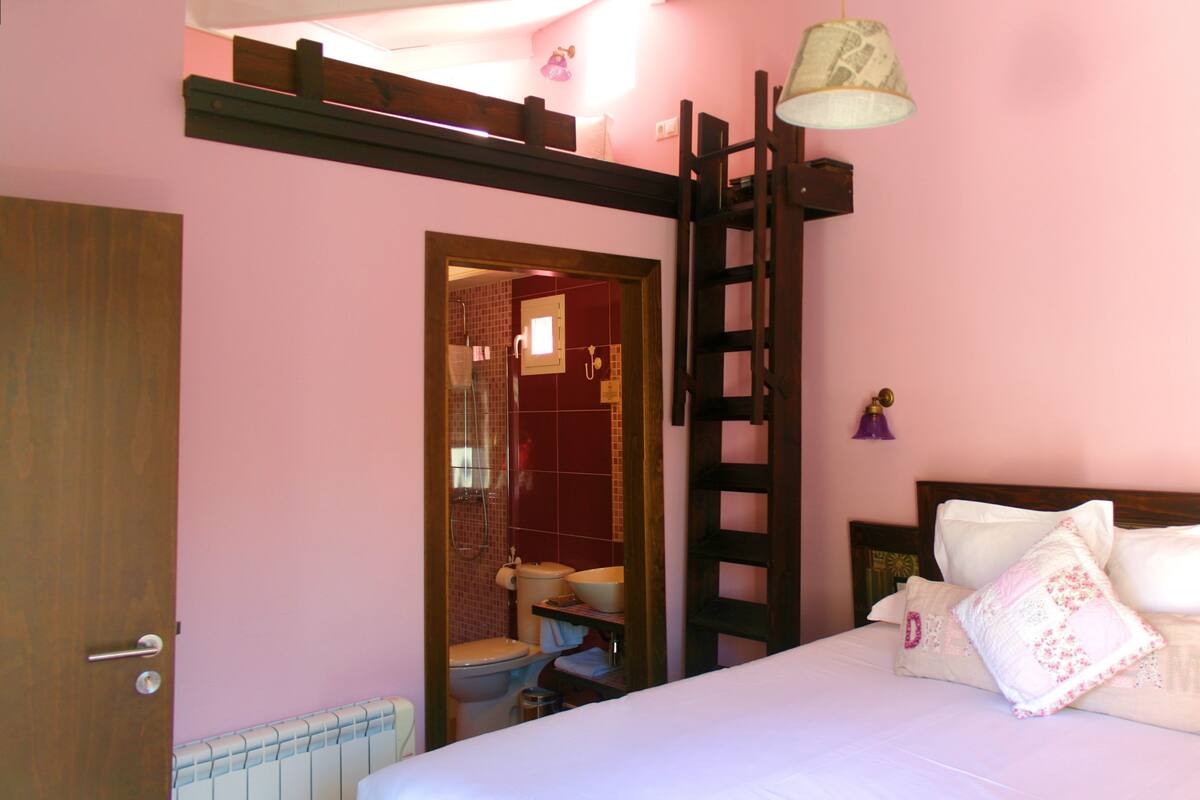 Un dormitorio con cama, una escalera de madera, un baño visible a través de una puerta abierta y una pared rosa.
