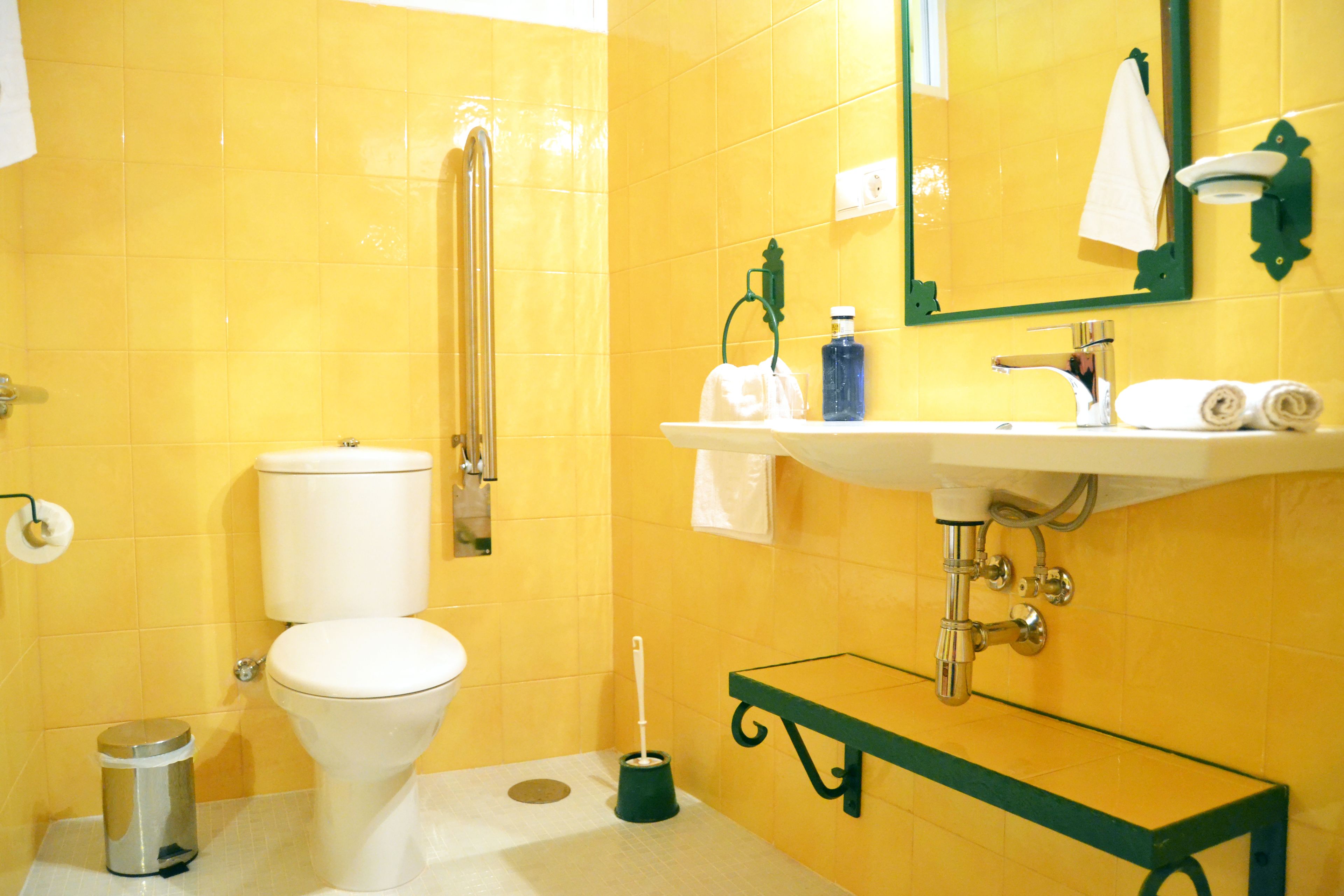 Un baño con paredes amarillas, un inodoro blanco, un lavabo de cristal con espejo y un banco verde.