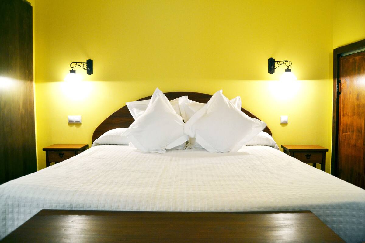 Una cama con almohadas blancas y cabecera de madera en una habitación con paredes amarillas y dos luces fijadas a la pared.