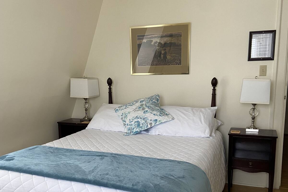 Un lit bien fait, avec une couette blanche matelassée et un plaid bleu. Il y a deux tables de chevet avec des lampes, un tableau encadré au-dessus du lit et une petite image accrochée au mur, à droite.