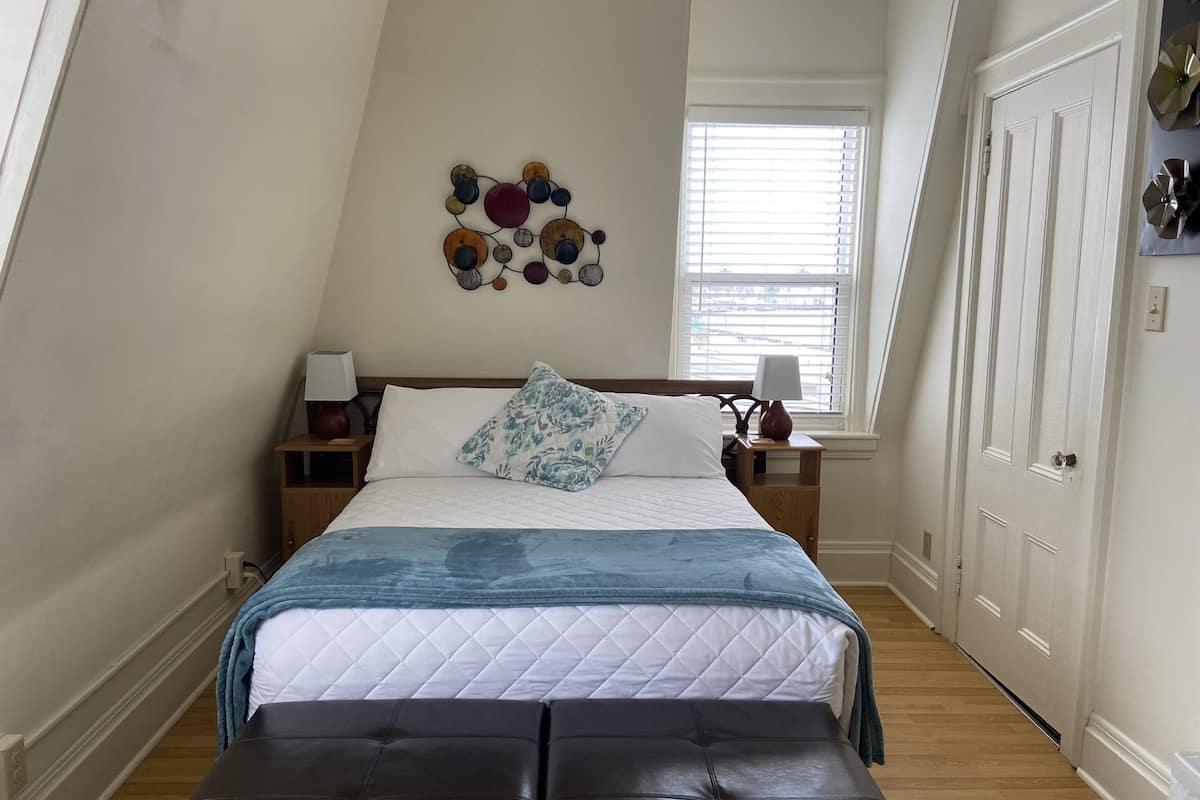Une chambre à coucher comprenant un lit, des tables de chevet, une lampe, un tableau décoratif et une fenêtre avec des stores.