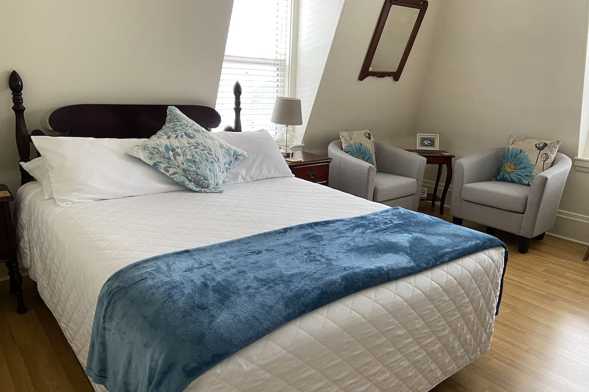 Un lit bien fait, avec une couverture bleue, une tête de lit en bois et deux chaises dans un coin.