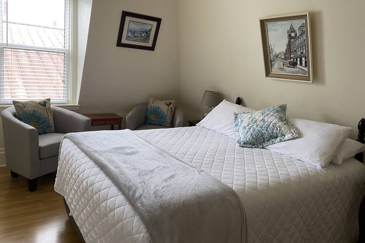 Une chambre à coucher avec un grand lit, deux chaises, une table de chevet et une fenêtre avec des stores.