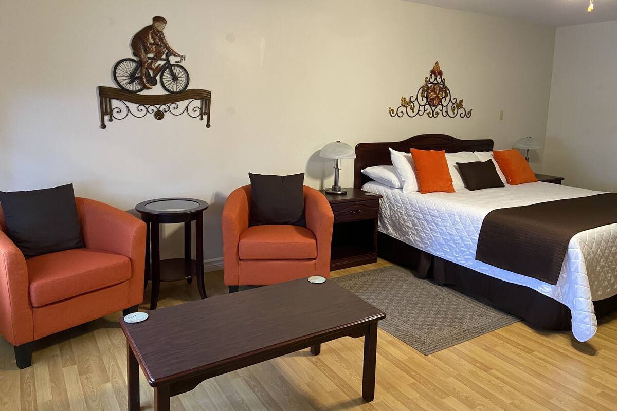 Une chambre d’hôtel comprenant un lit, deux fauteuils, une petite table, une table de chevet, une lampe et des œuvres d’art décoratives accrochées au mur.
