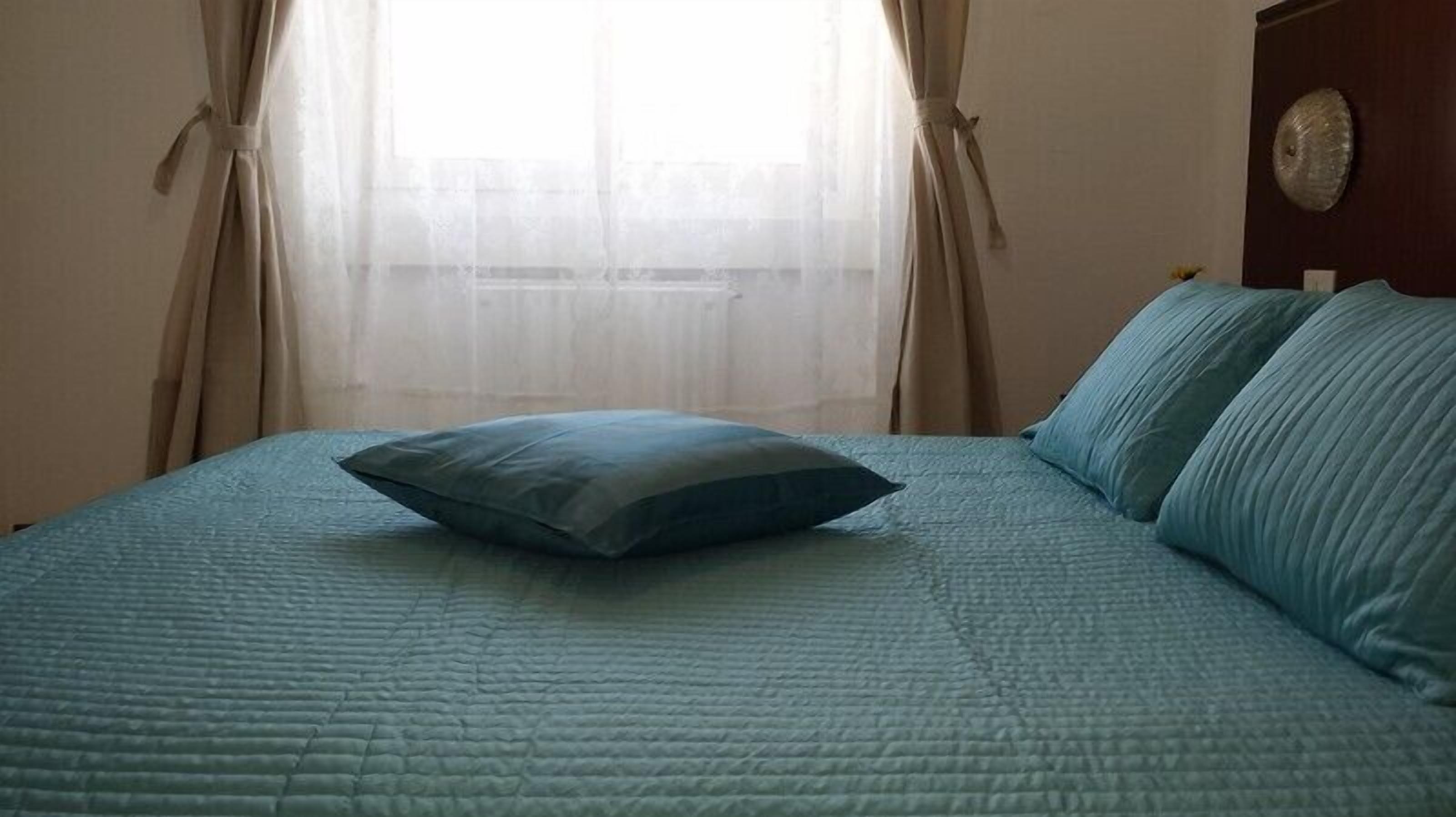 A bed with a blue quilt and pillows, a window with sheer curtains, and a wooden headboard.