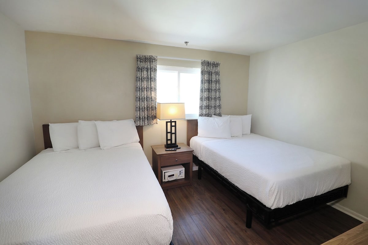 A hotel room with two single beds, a nightstand with a lamp, and a small table with a digital clock.