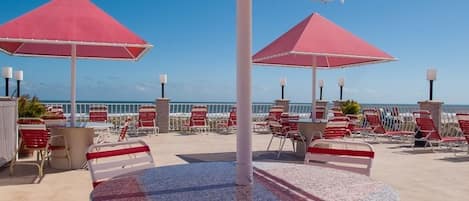 A rooftop terrace with red umbrellas, lounge chairs, and a view of the ocean.