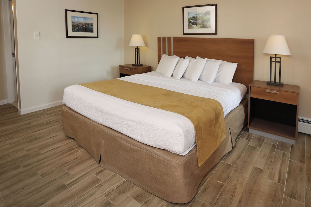 A neatly made bed with a brown headboard, white bedding, and a brown throw blanket. There are two bedside tables with lamps, and the room has wooden flooring.