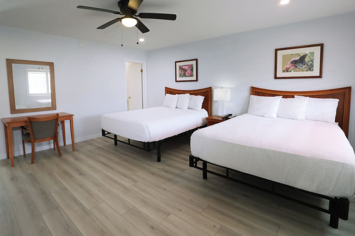 A hotel room with two beds, a desk, a chair, a ceiling fan, and two framed pictures on the walls.