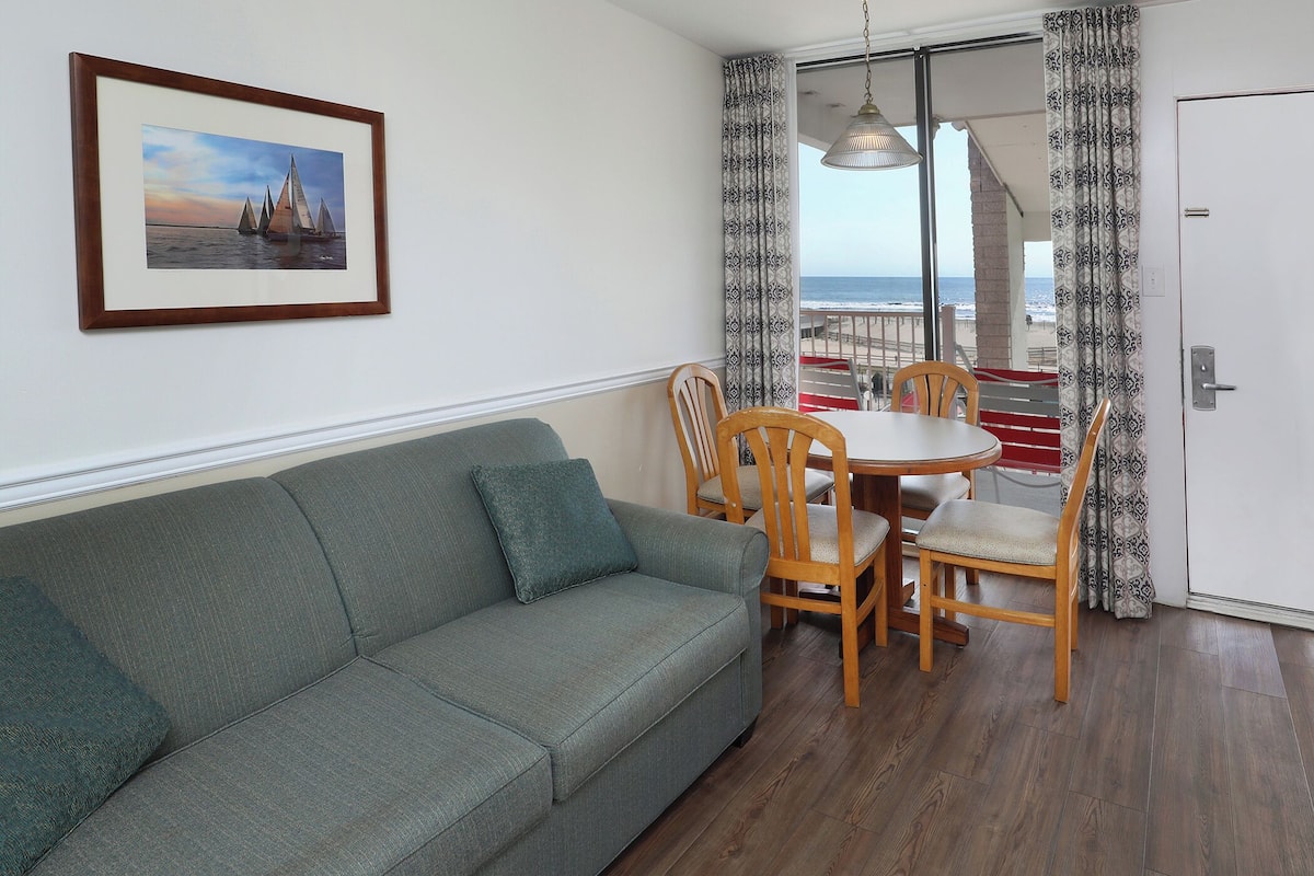 A living room with a sofa, a dining table with chairs, a framed picture on the wall, and a view of the sea through a glass door.