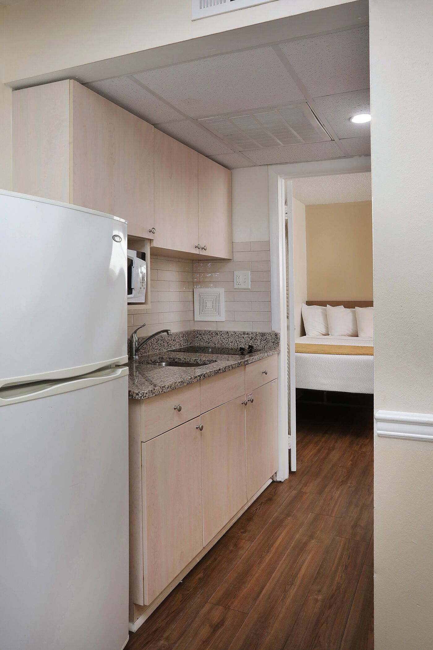 Cocina compacta con refrigerador, pileta y alacenas de madera. Se ve una cama en la habitación contigua.