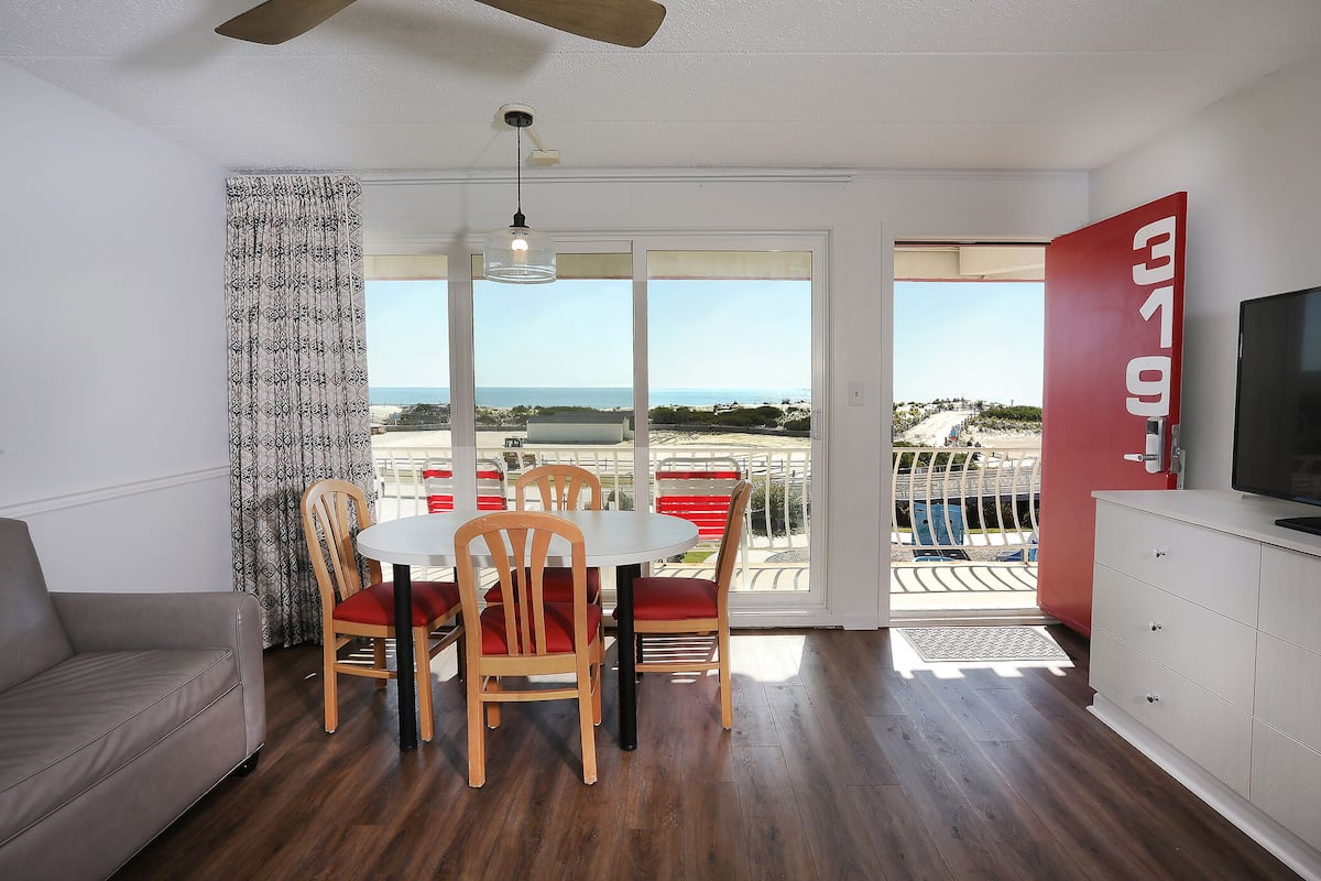 A room with a dining table, chairs, a sofa, a TV, and a balcony with a view of the beach.