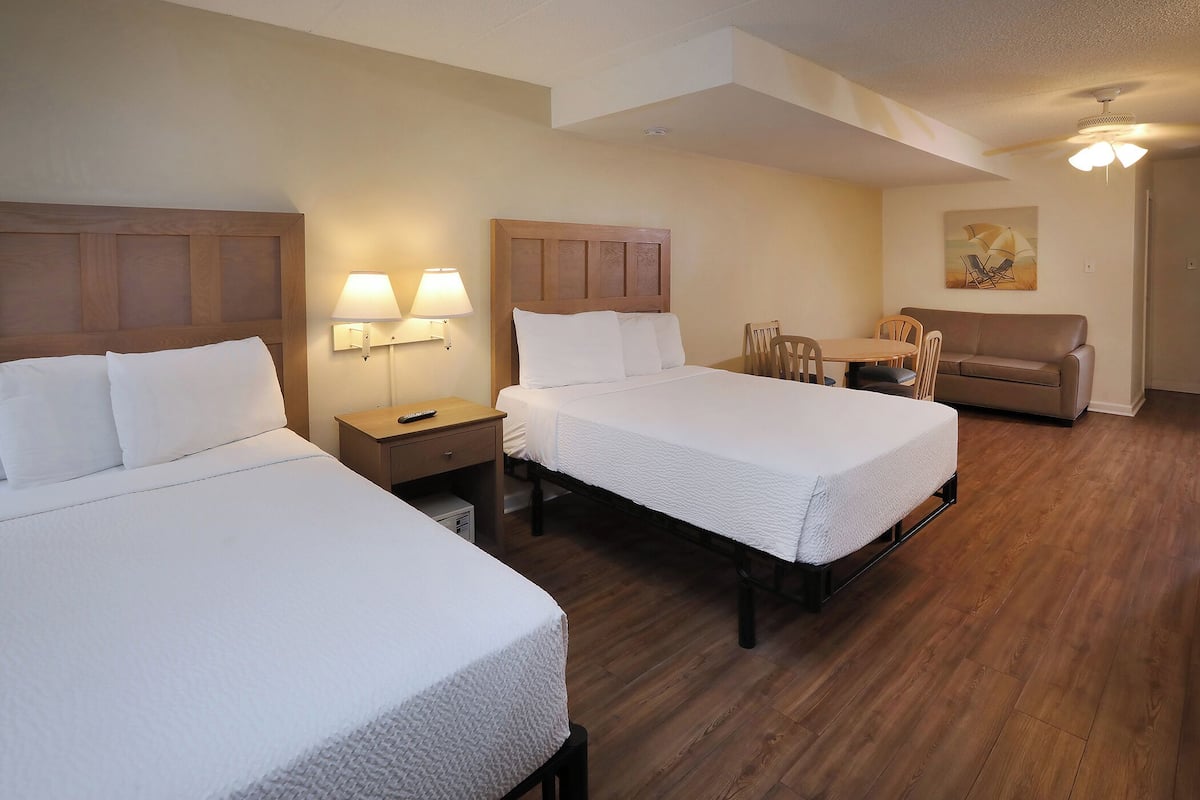 A hotel room with two beds, a sofa, a dining table with chairs, and a ceiling fan.