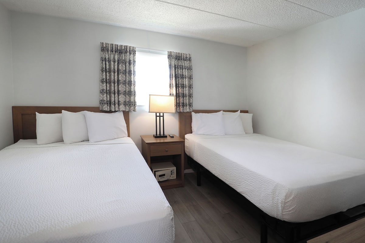 A hotel room with two single beds, a nightstand with a lamp, and a window with patterned curtains.