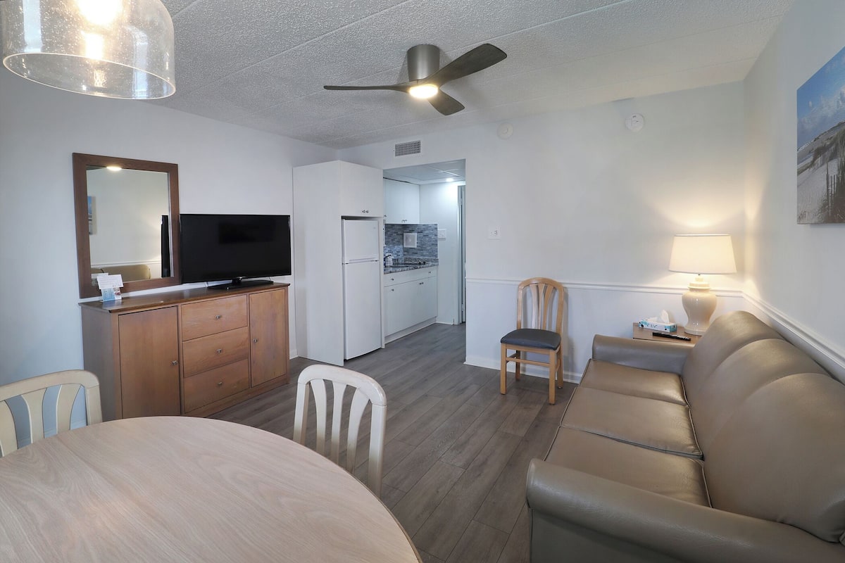 A compact living space with a dining area, a sofa, a TV, a kitchen, and a ceiling fan.