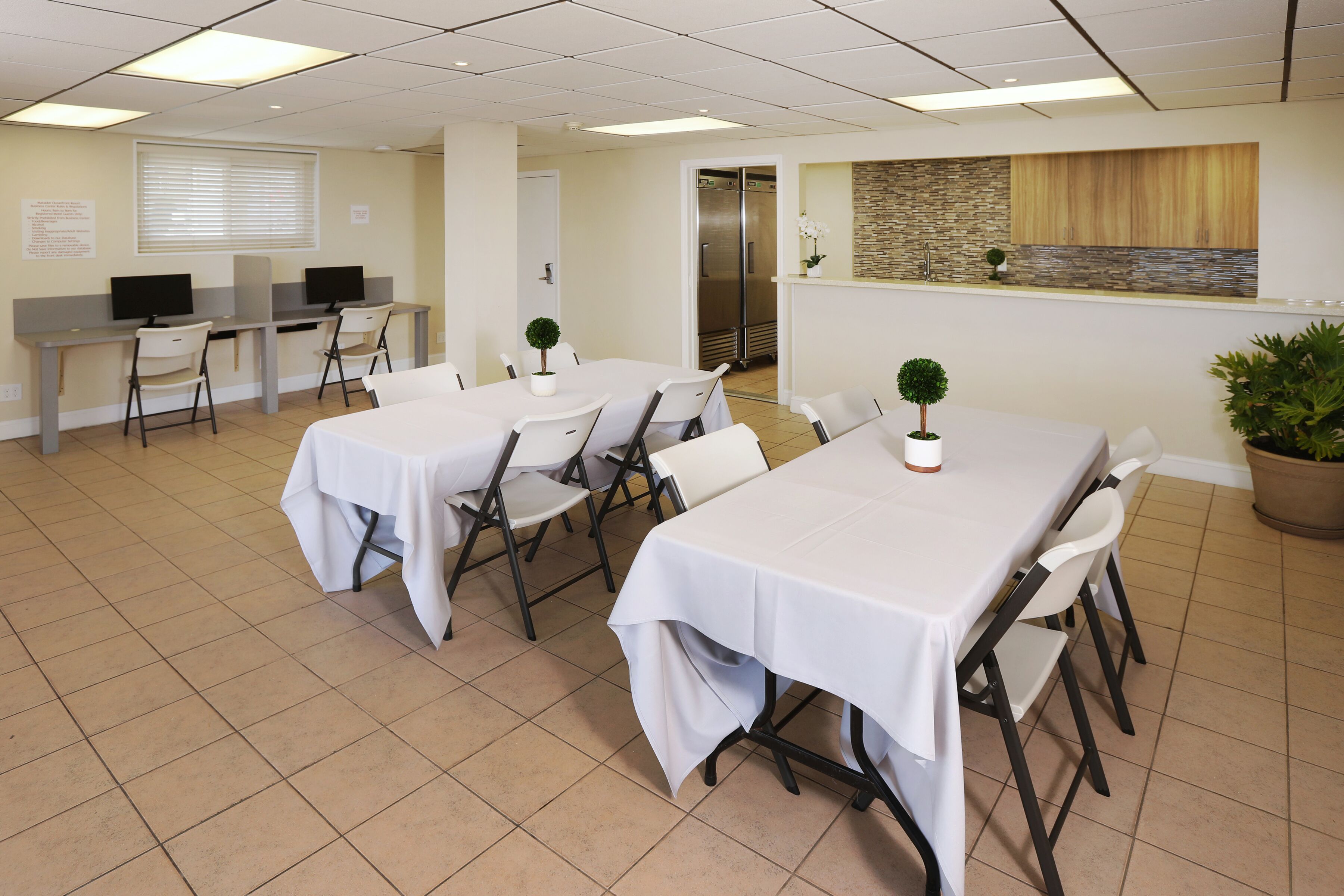 A conference room with two long tables, chairs, a small potted plant, a refrigerator, and a stone accent wall.