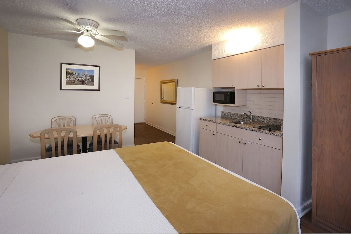 A compact room with a bed, a dining table with chairs, a kitchenette with a microwave, and a ceiling fan.