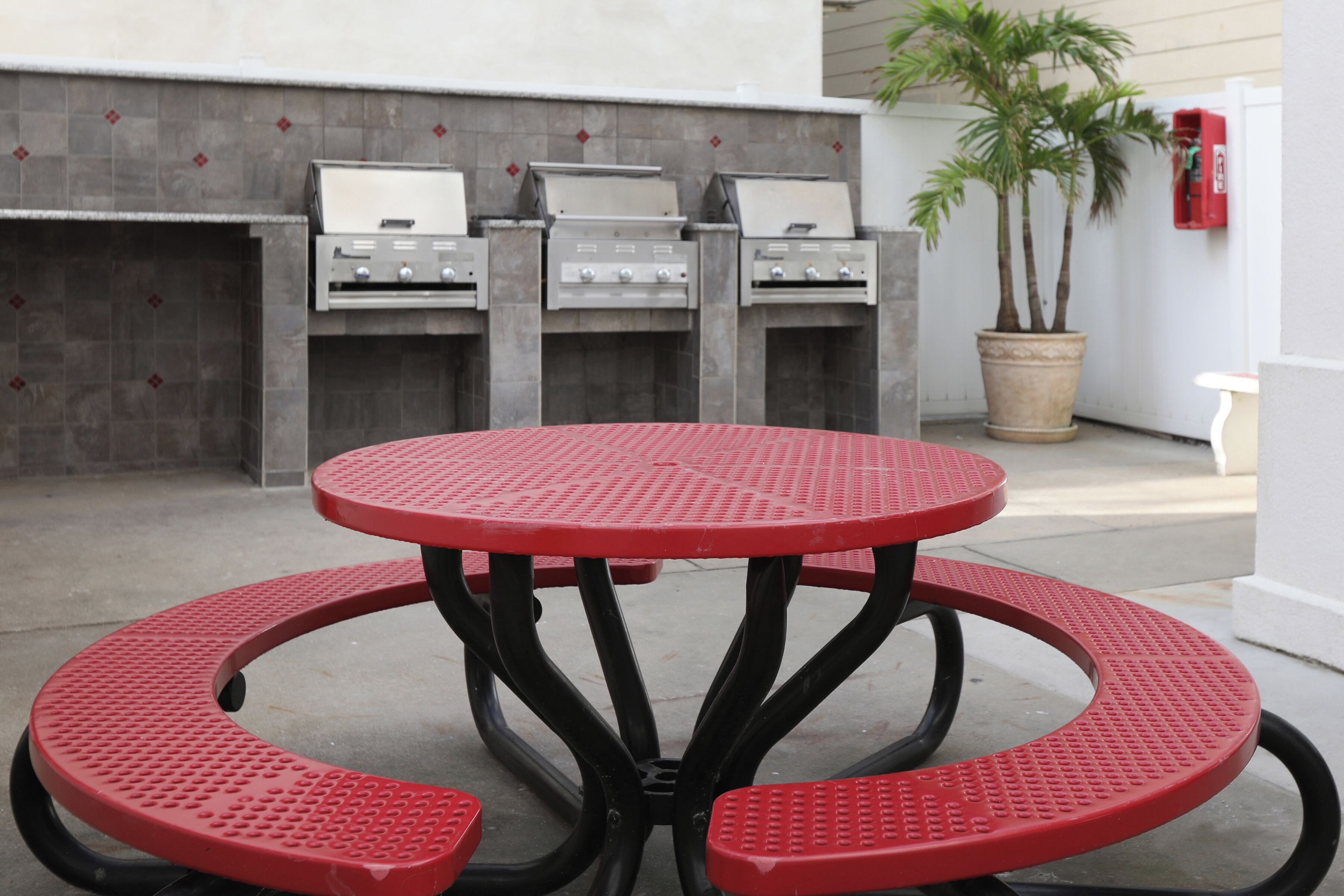 Une table de pique-nique en métal rouge, de forme circulaire, placée devant un décor composé de trois barbecues et d’une plante en pot.