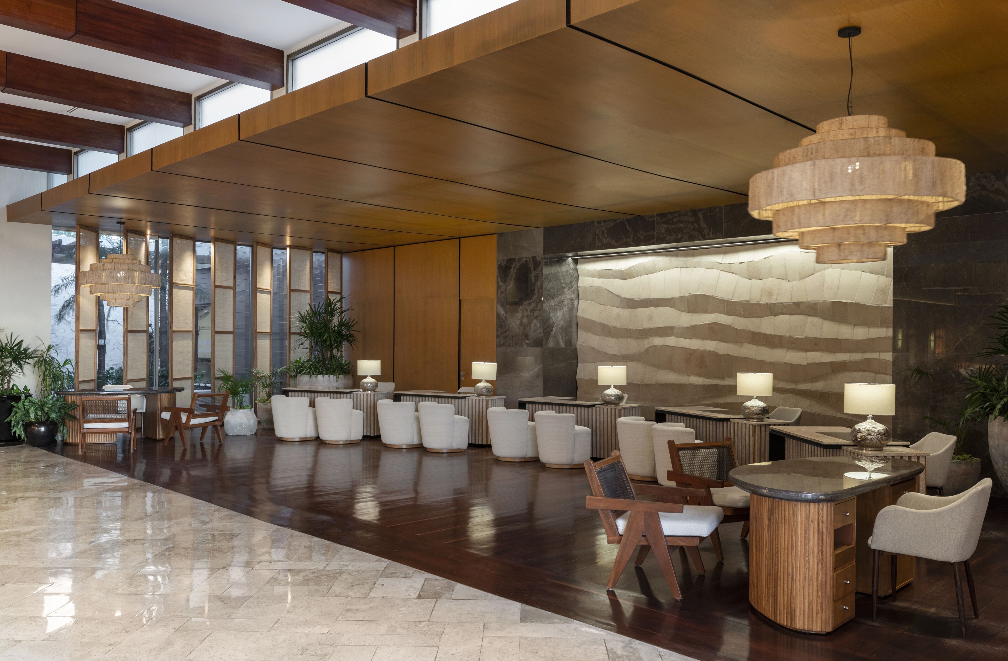 A spacious lobby with a high ceiling, wooden paneling, and a large stone wall. There are several seating areas with tables and chairs, and multiple lamps providing lighting.