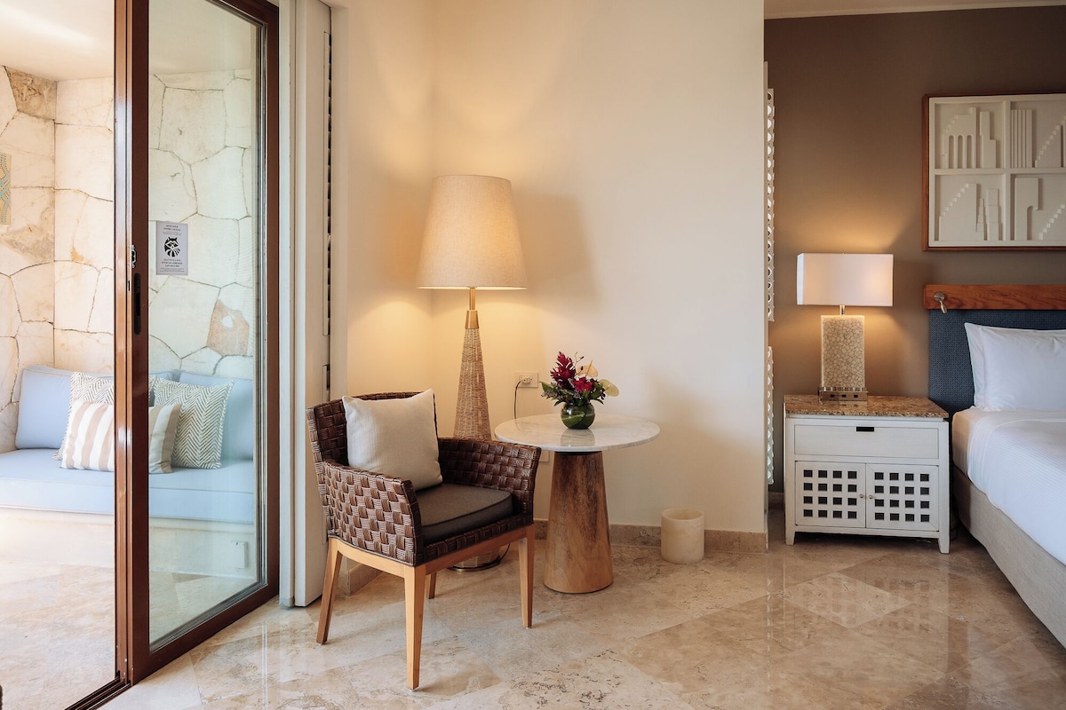 A hotel room with a glass door, a wicker chair, a small round table with a lamp, a bedside table with a lamp, and a bed.