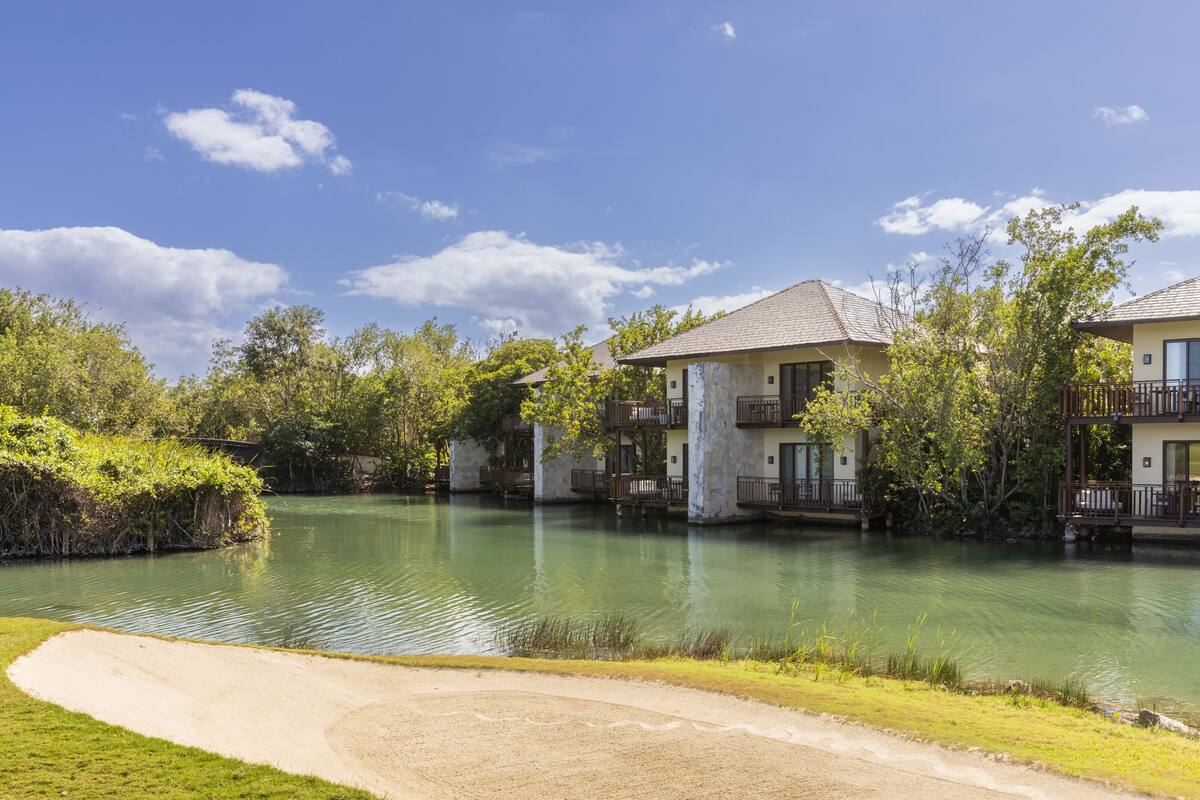 A lakeside hotel with multiple-story buildings, a sandy pathway, and lush greenery.