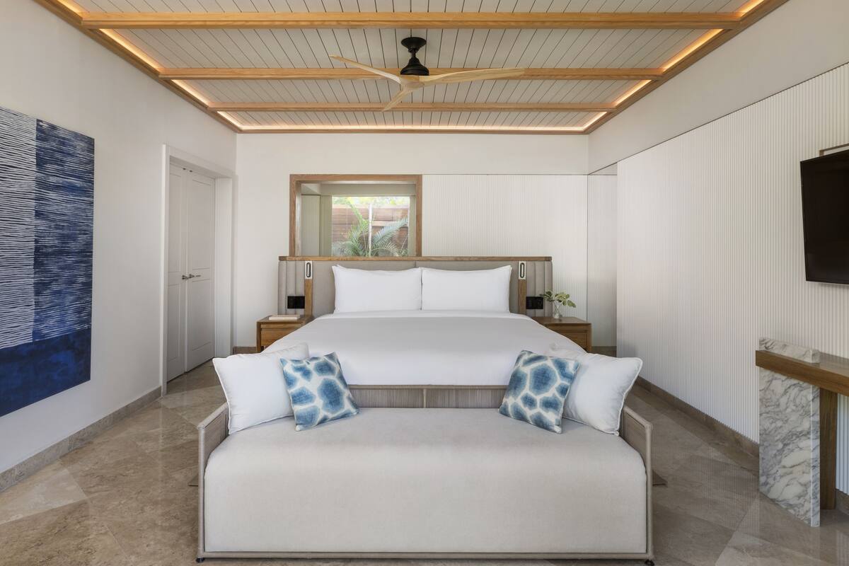 A modern bedroom with a large bed, a sofa, a ceiling fan, and a TV on the wall.
