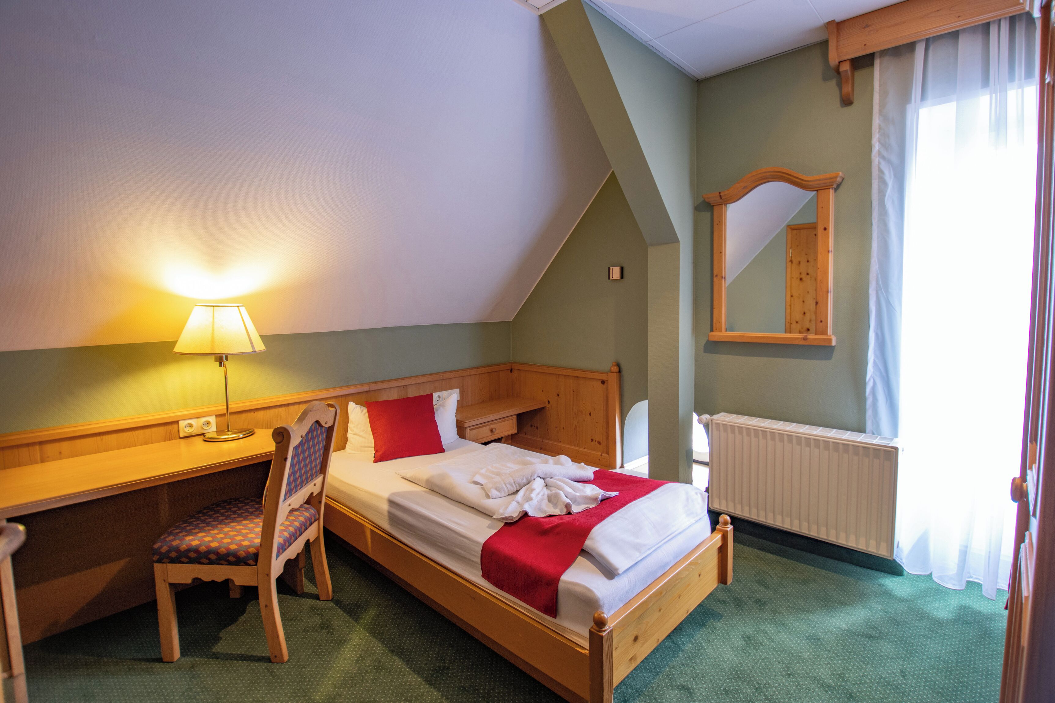Habitación de hotel con cama de madera, escritorio, silla, lámpara, espejo y radiador.
