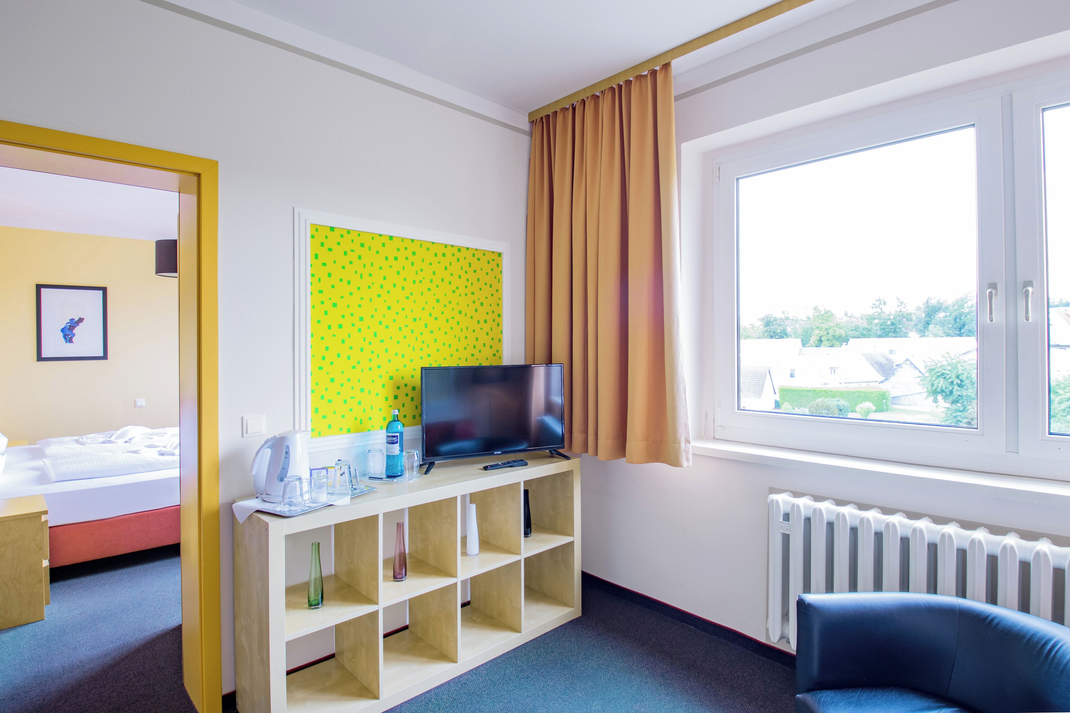 A hotel room with a bed, a TV on a wooden stand, a window with curtains, and a view of greenery outside.