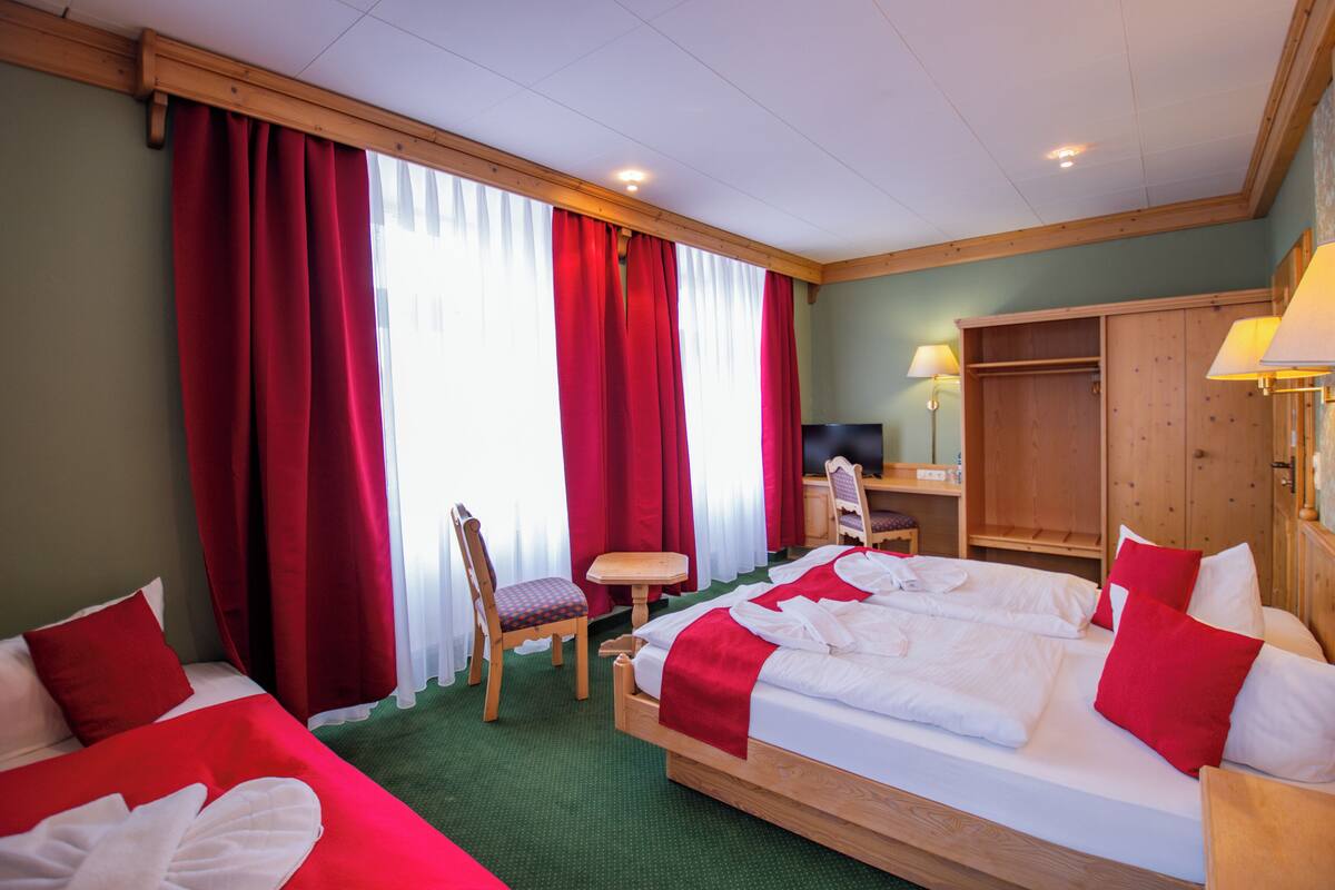Ein Hotelzimmer mit zwei Betten, roten Vorhängen, einem Holztisch, einem Stuhl und einem Kleiderschrank.
