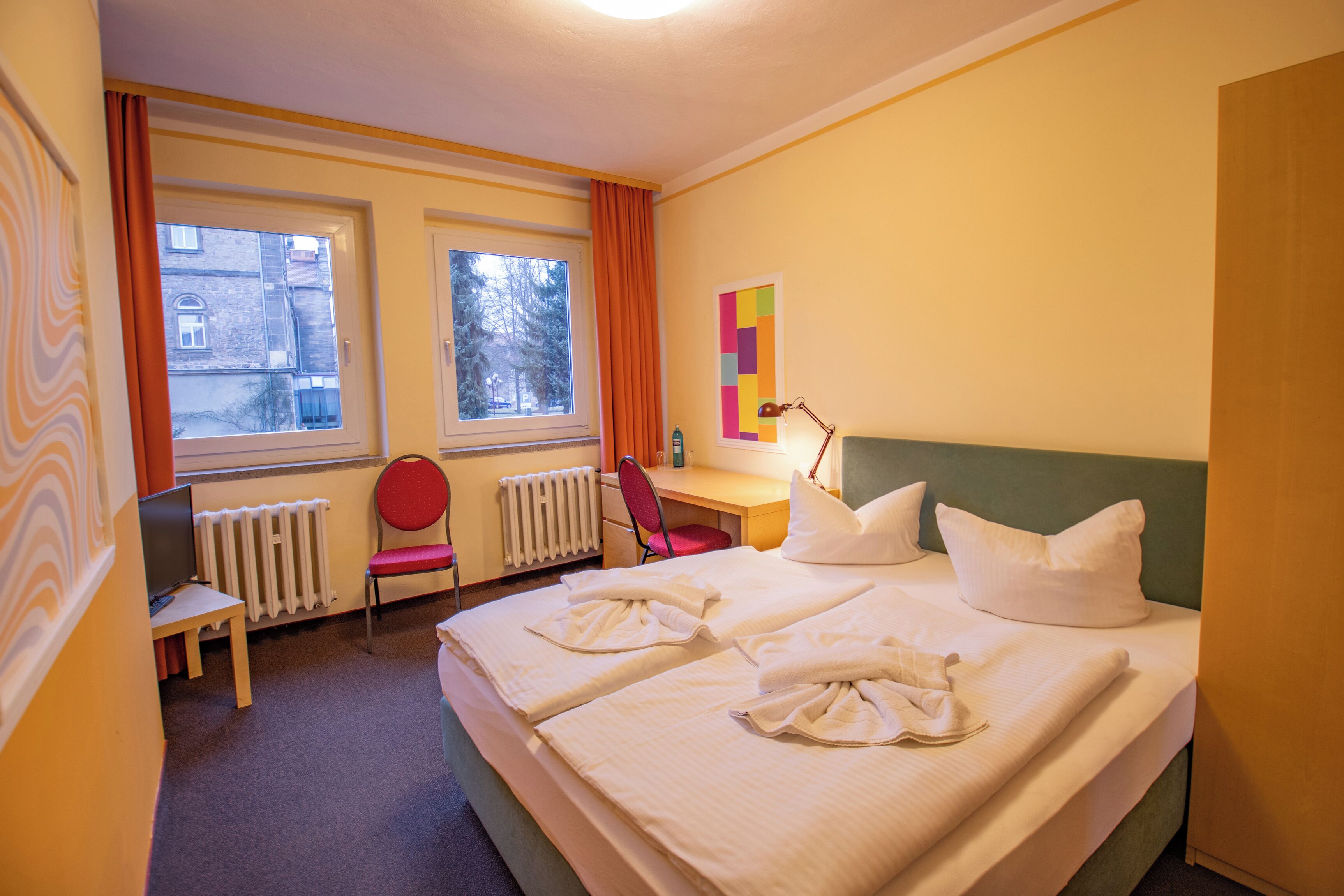 A hotel room with two beds, a desk, a chair, a radiator, and a window with curtains.