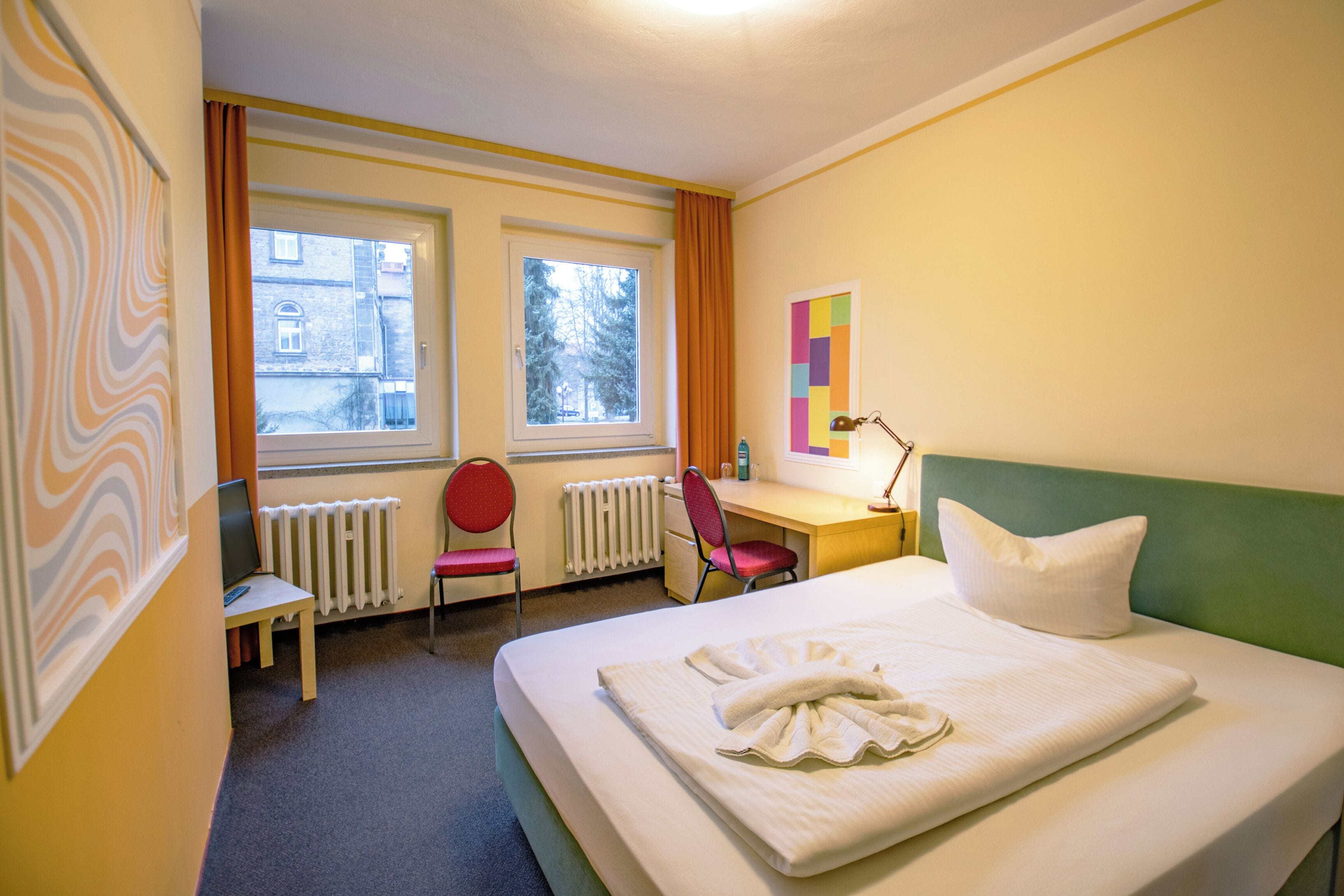 Une chambre d’hôtel avec un lit, un bureau, une chaise et deux fenêtres.