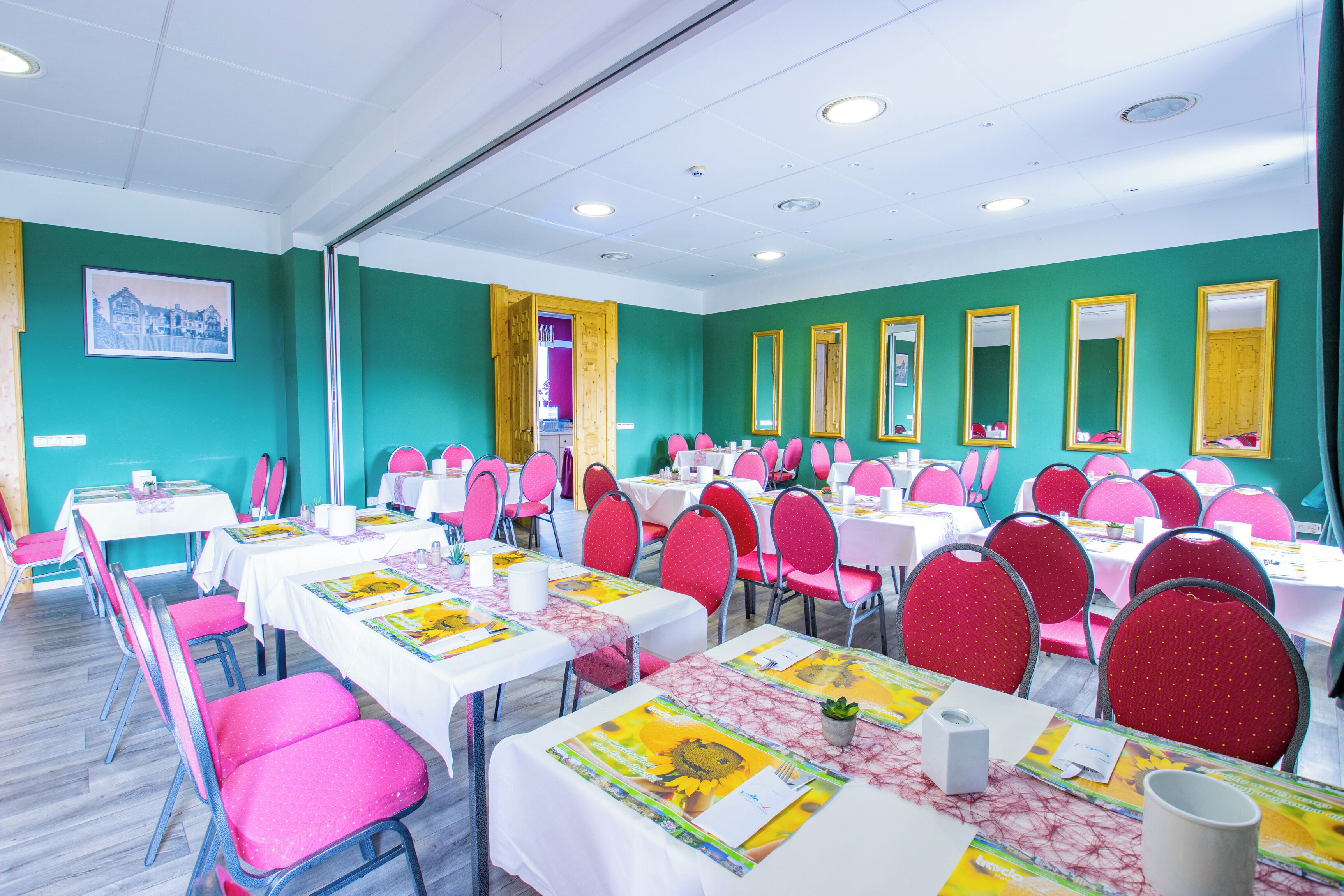 Un salón de conferencias con varias mesas y sillas, paredes color turquesa y un ventanal grande.
