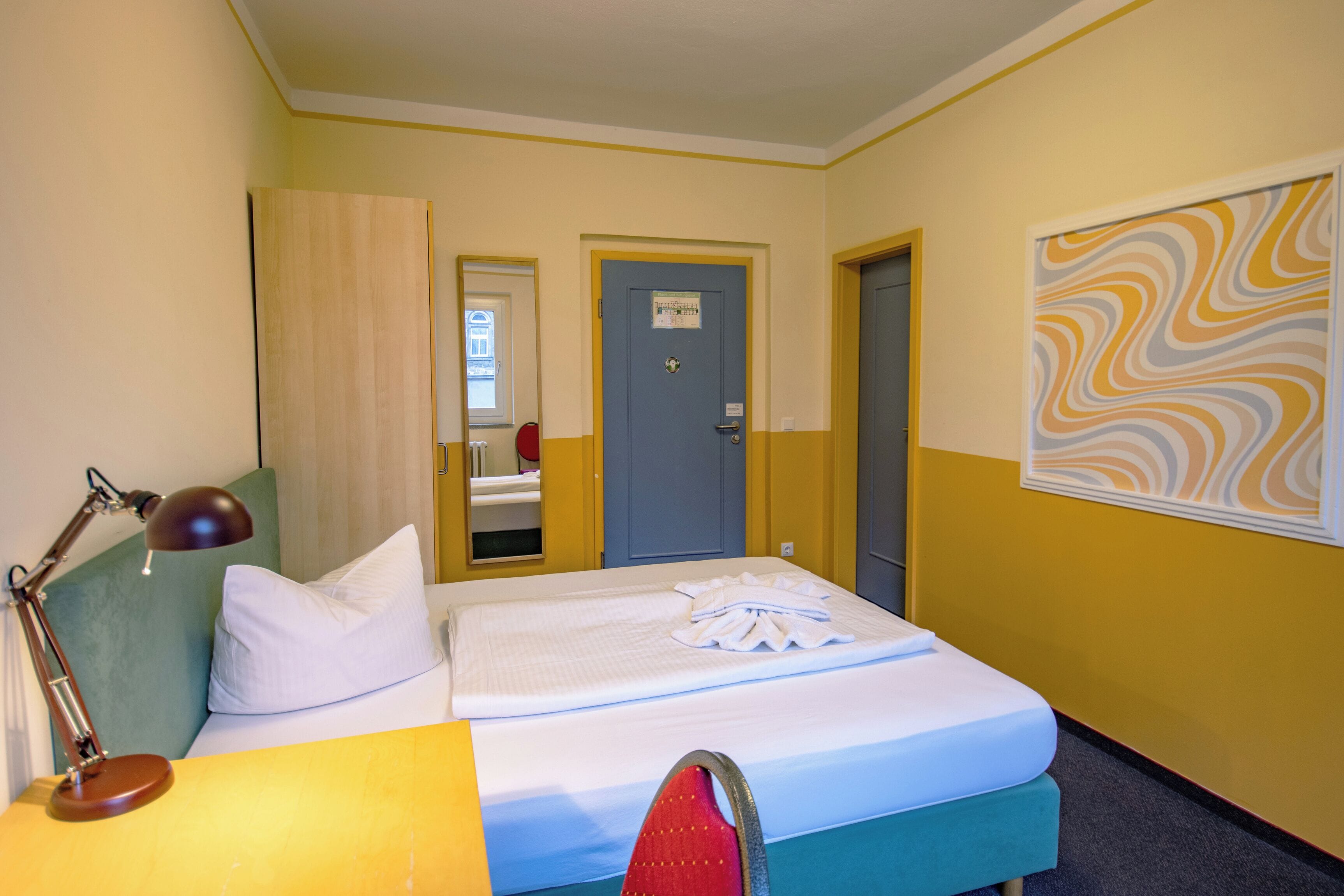 A hotel room with a bed, a desk, a lamp, and a door.