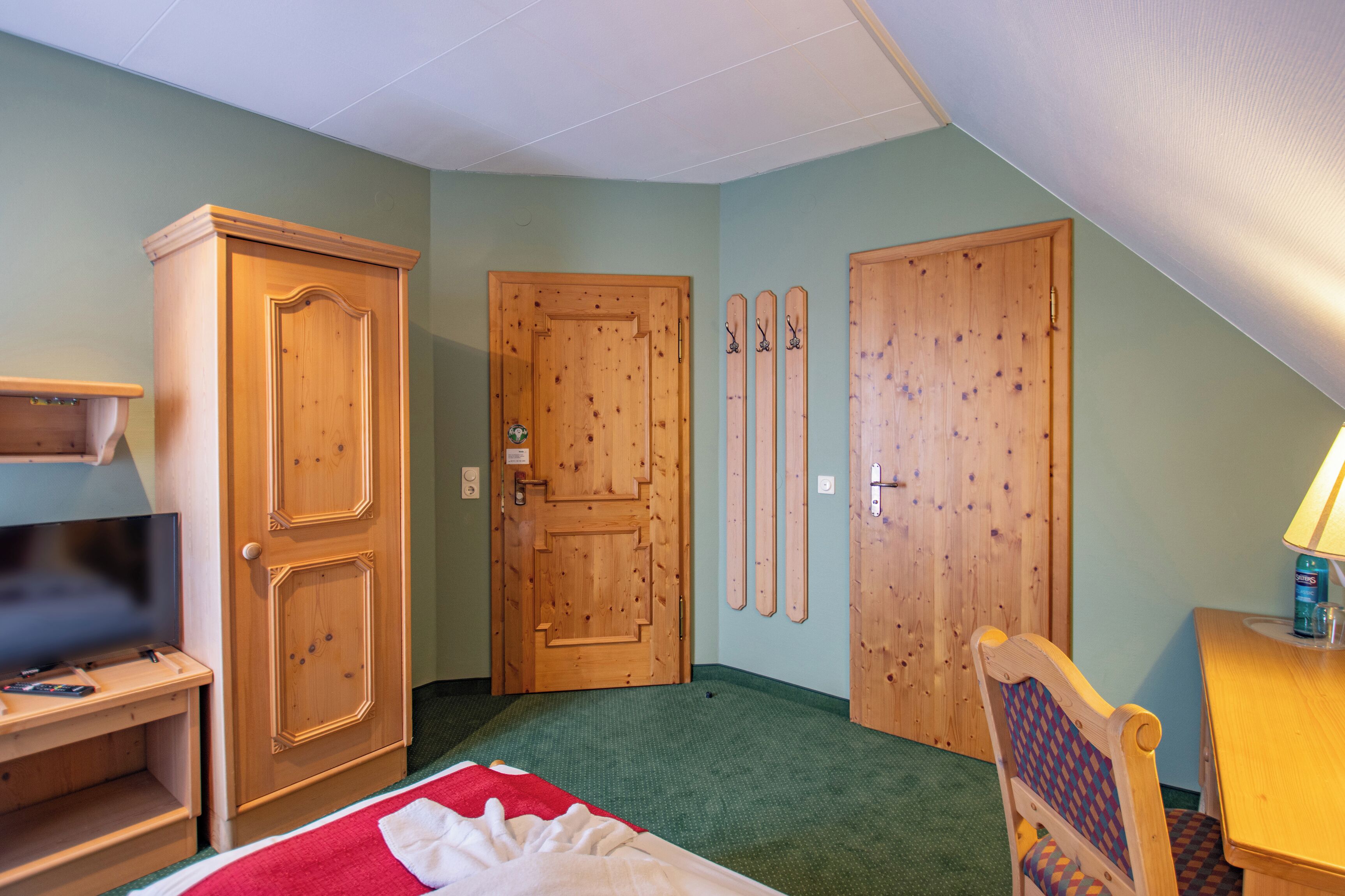 Una stanza con un letto, un armadio in legno, una televisione, una scrivania e una sedia. Due porte in legno e un paio di sci appesi al muro.