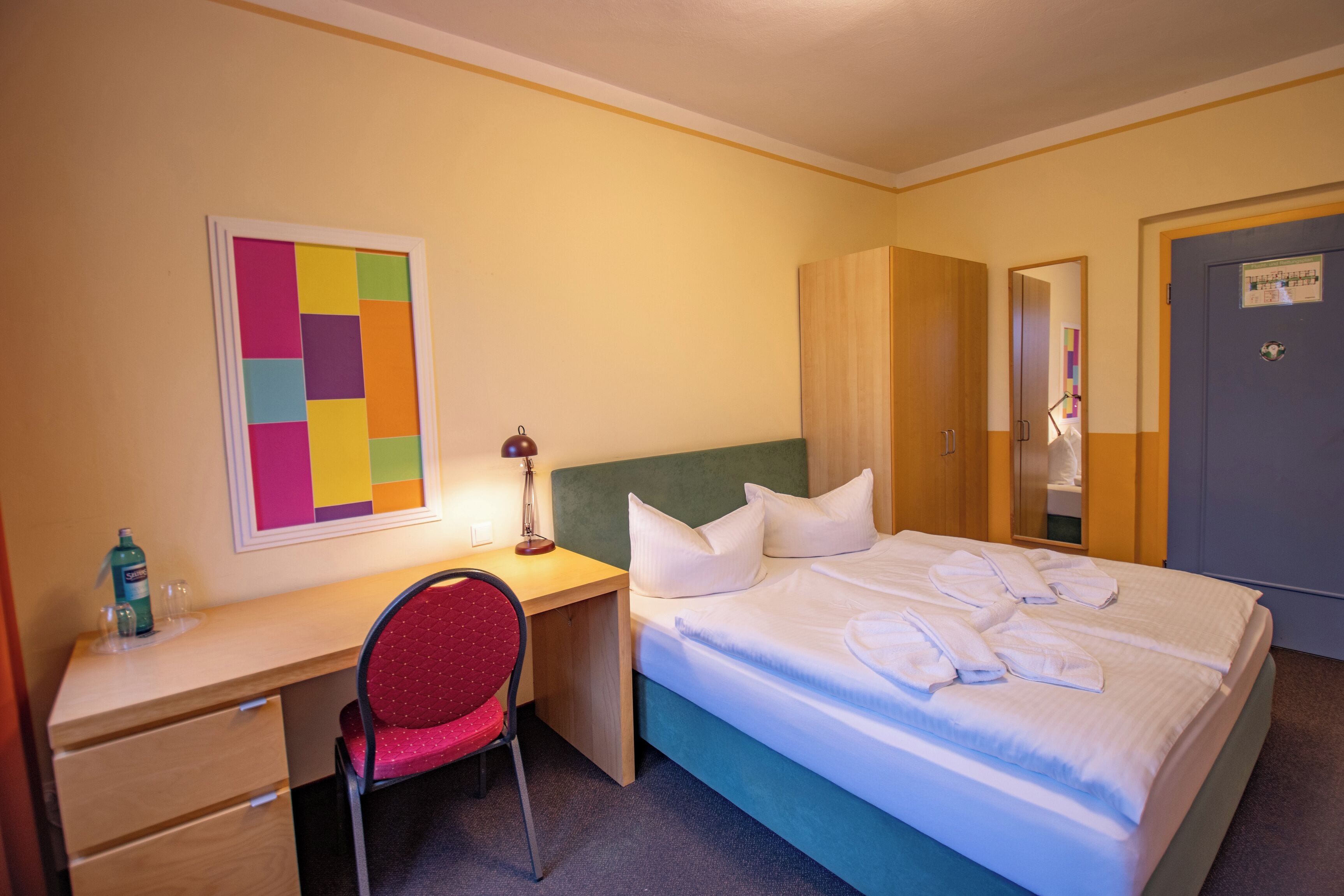 A hotel room with a bed, a desk, a chair, and a colorful artwork on the wall.