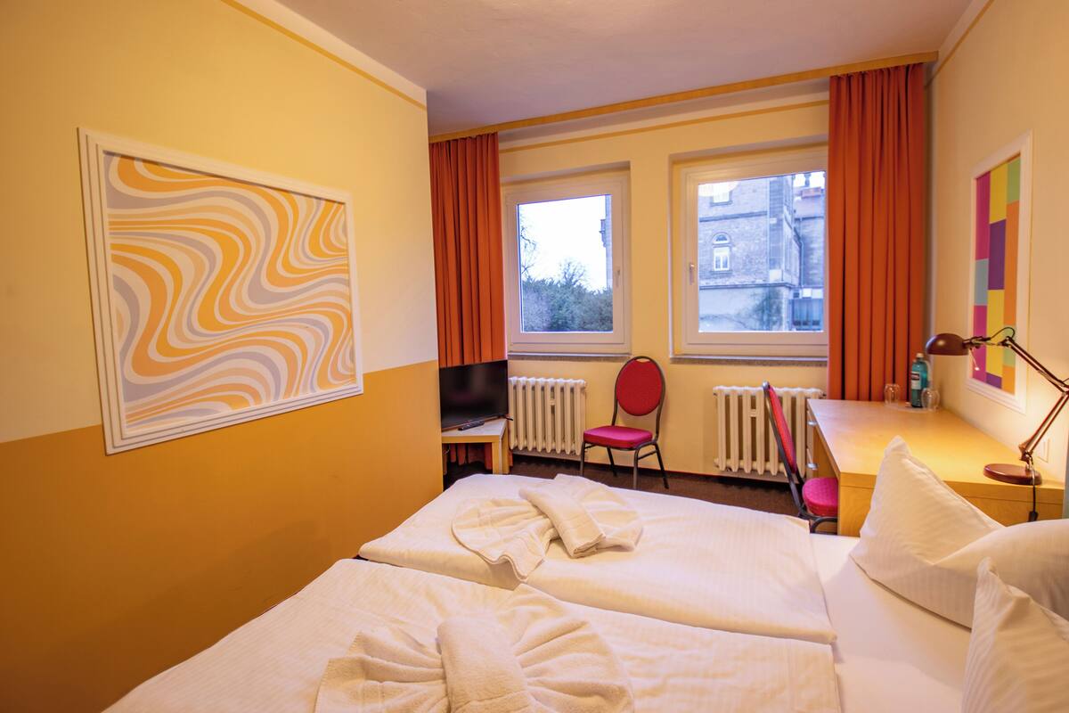 Ein Hotelzimmer mit zwei Betten, einem Fernseher, einem Sessel und einem Fenster mit Vorhängen.