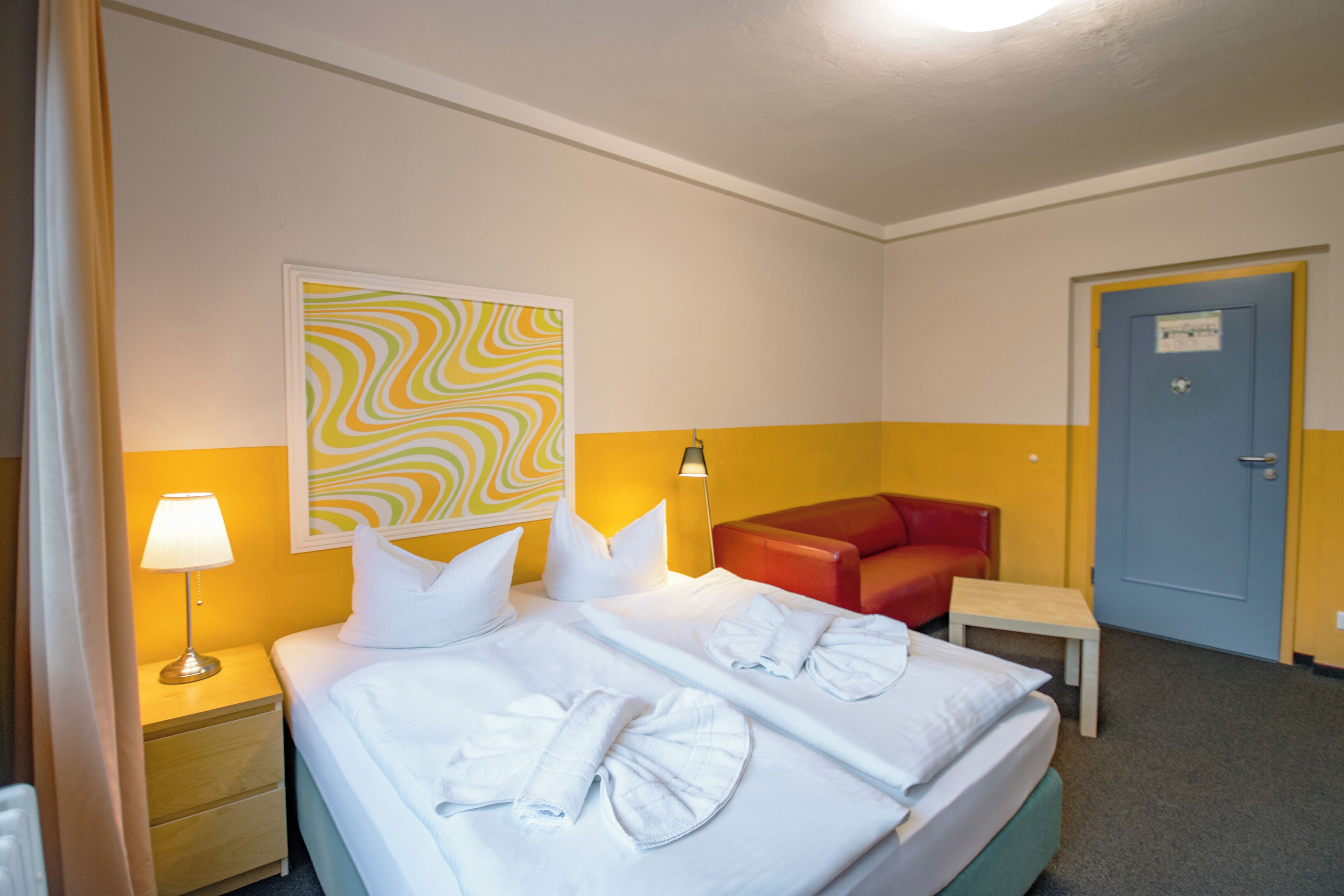 A hotel room with a bed, a nightstand, a lamp, a red sofa, a small table, and a door.