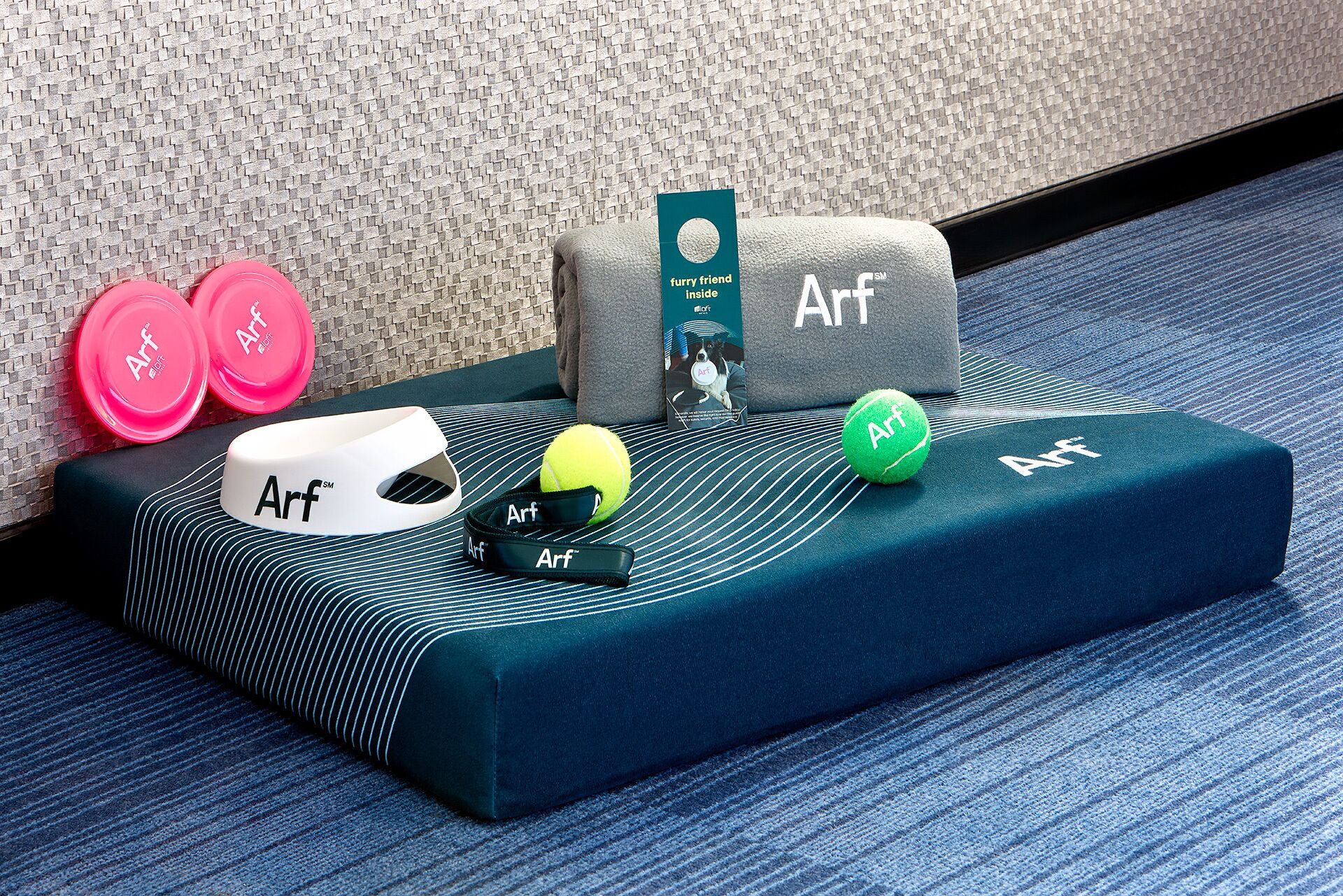 A hotel room with a bed, pillows, a blanket, a pair of slippers, a tennis ball, and a book.