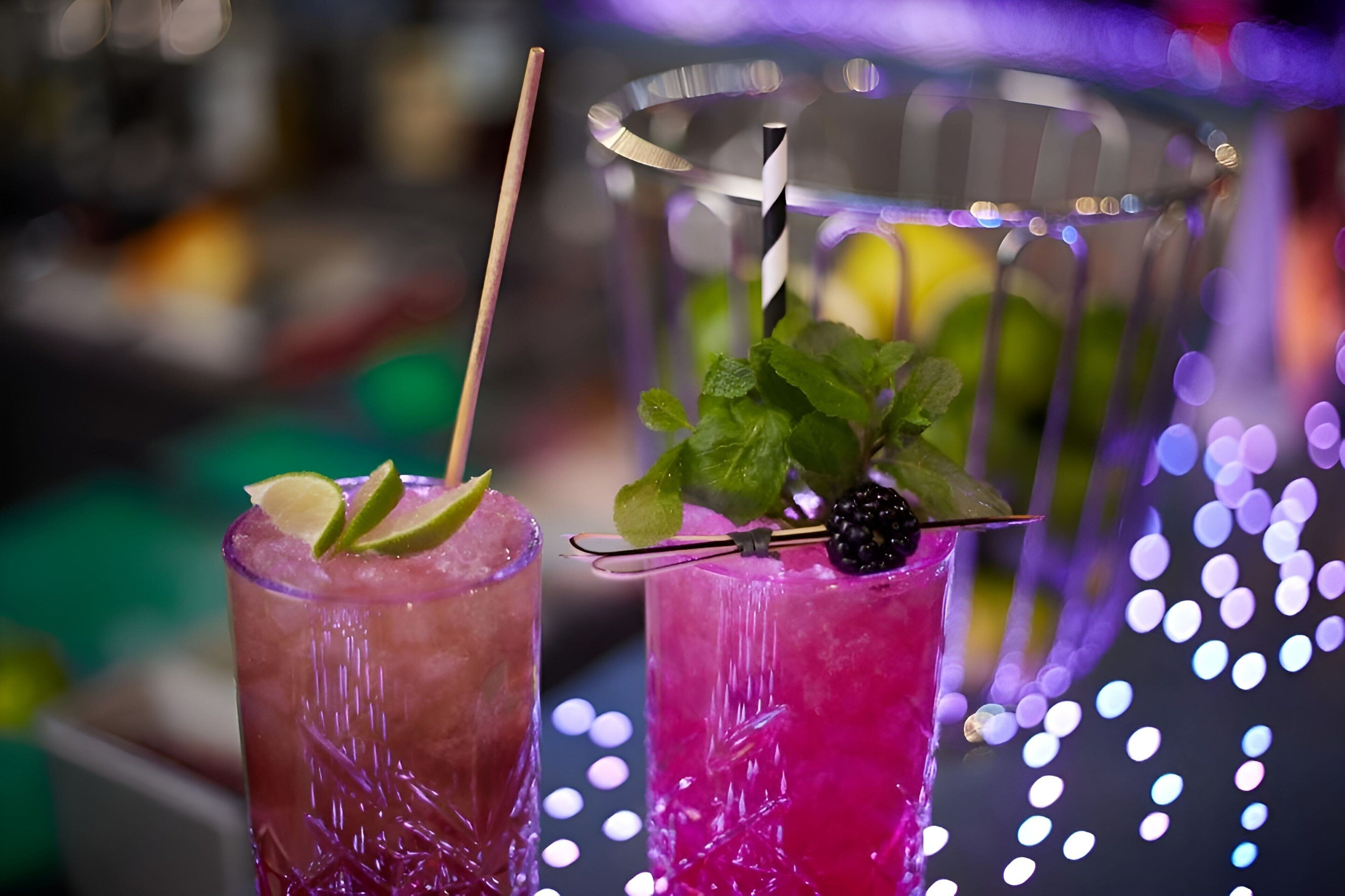 Two glasses of pink cocktails with lime and mint garnishes, one with a straw and one with a blackberry.