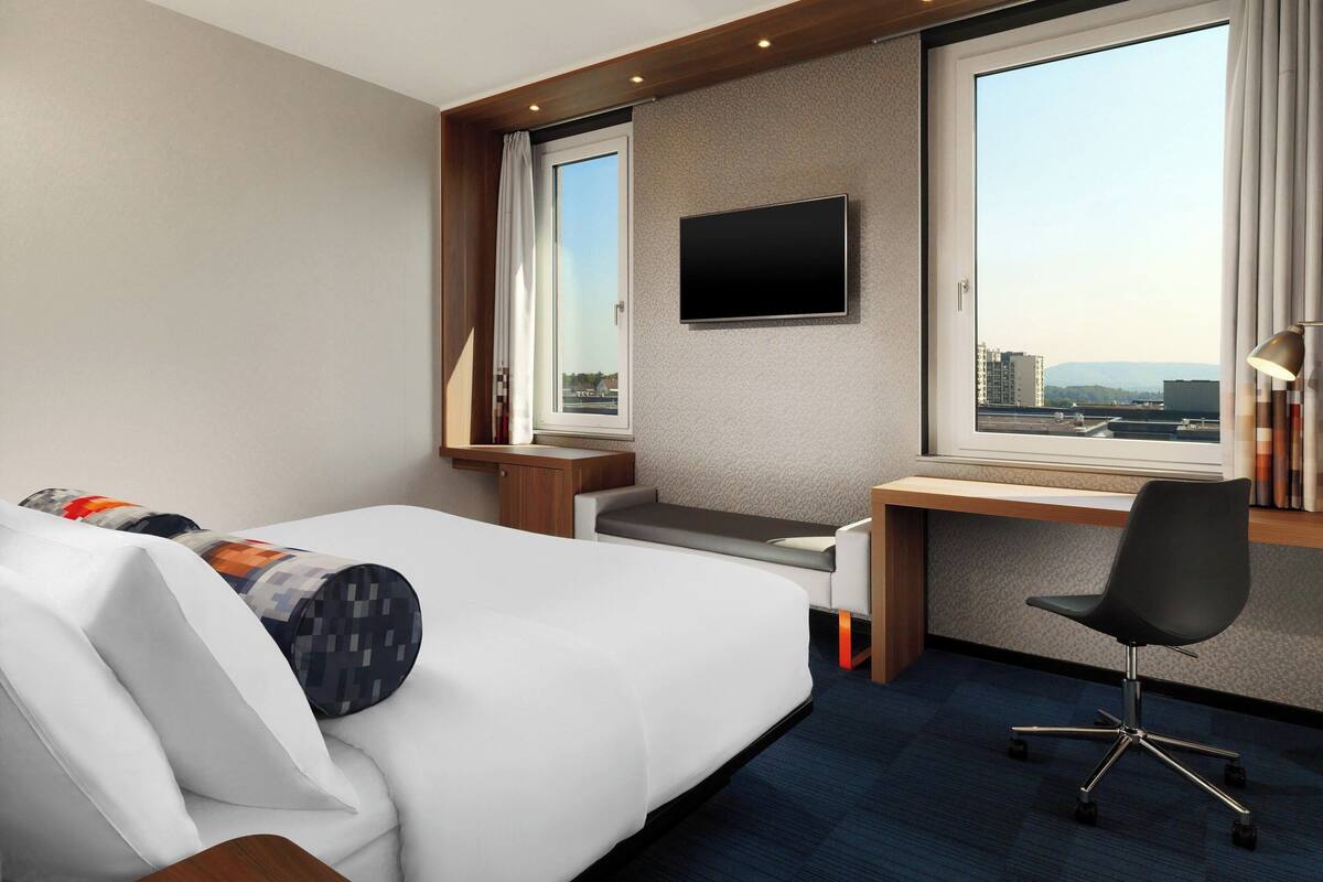A hotel room with a bed, a desk, a chair, a TV, and a large window with a city view.
