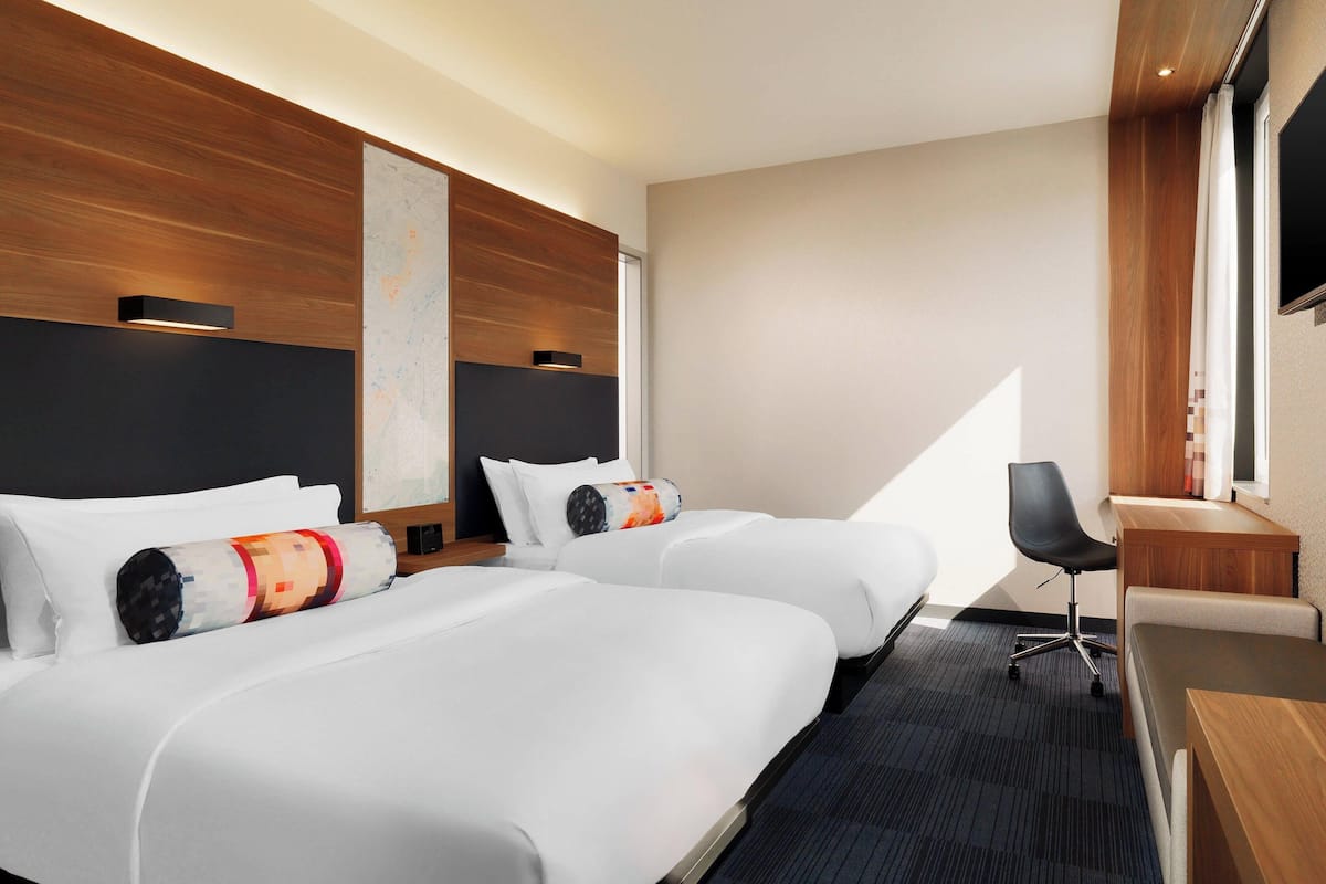 A hotel room with two beds, a desk, a chair, and a large window.